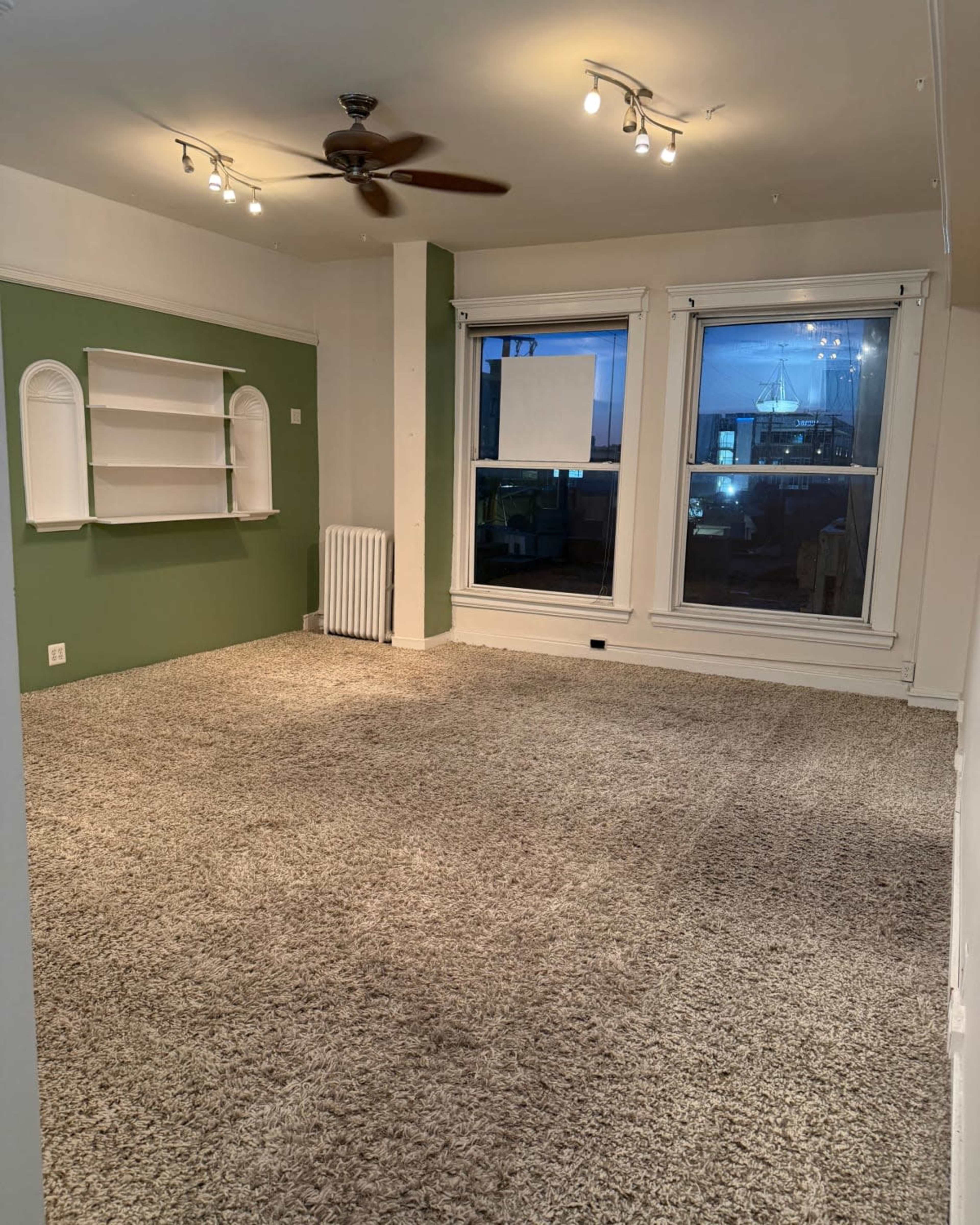 The image shows a spacious, empty room with a green accent wall, a ceiling fan, and large windows revealing a view outside.