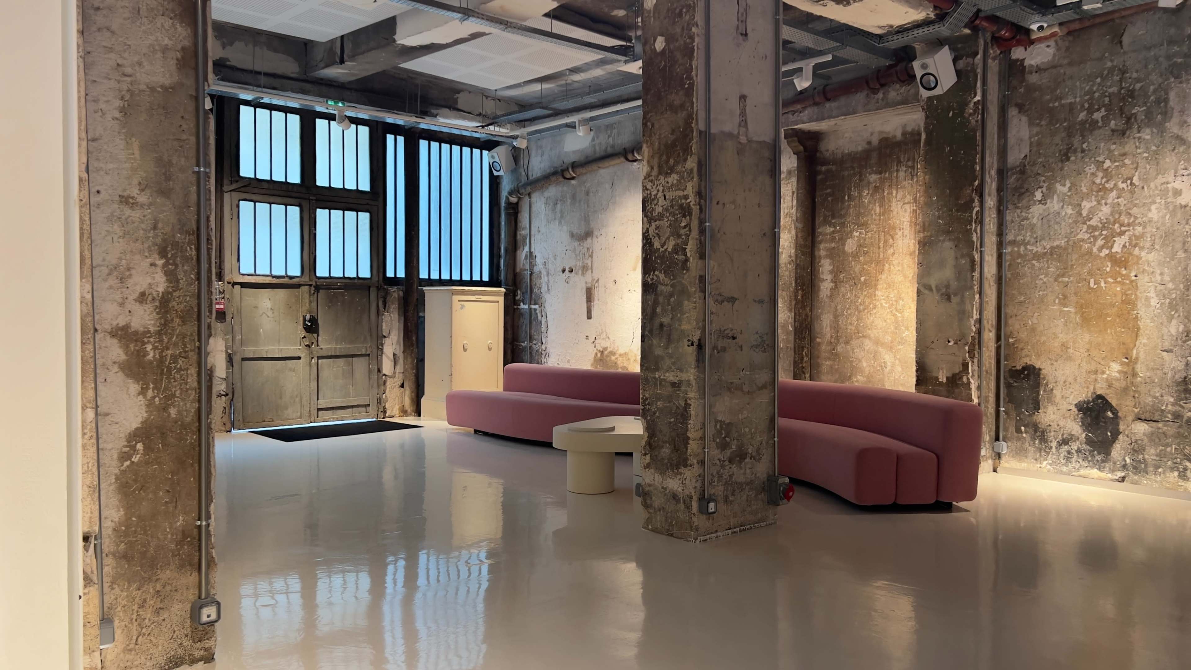 The image shows a spacious interior with a pink curved sofa, a small round table, and exposed concrete pillars, illuminated by natural light coming through large windows.
