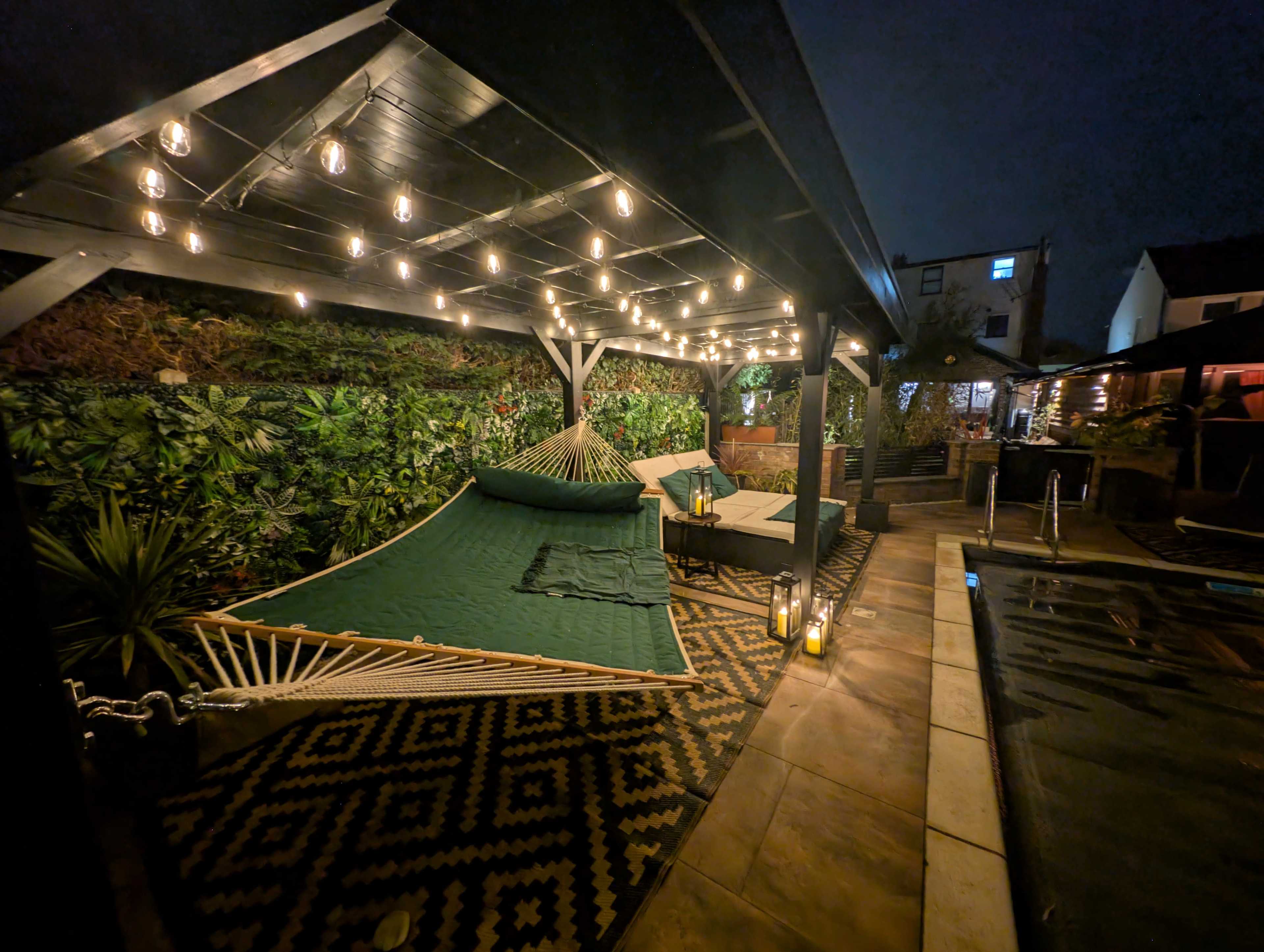 The image shows a backyard patio area with a hammock, lounge chairs, and string lights illuminating the space.