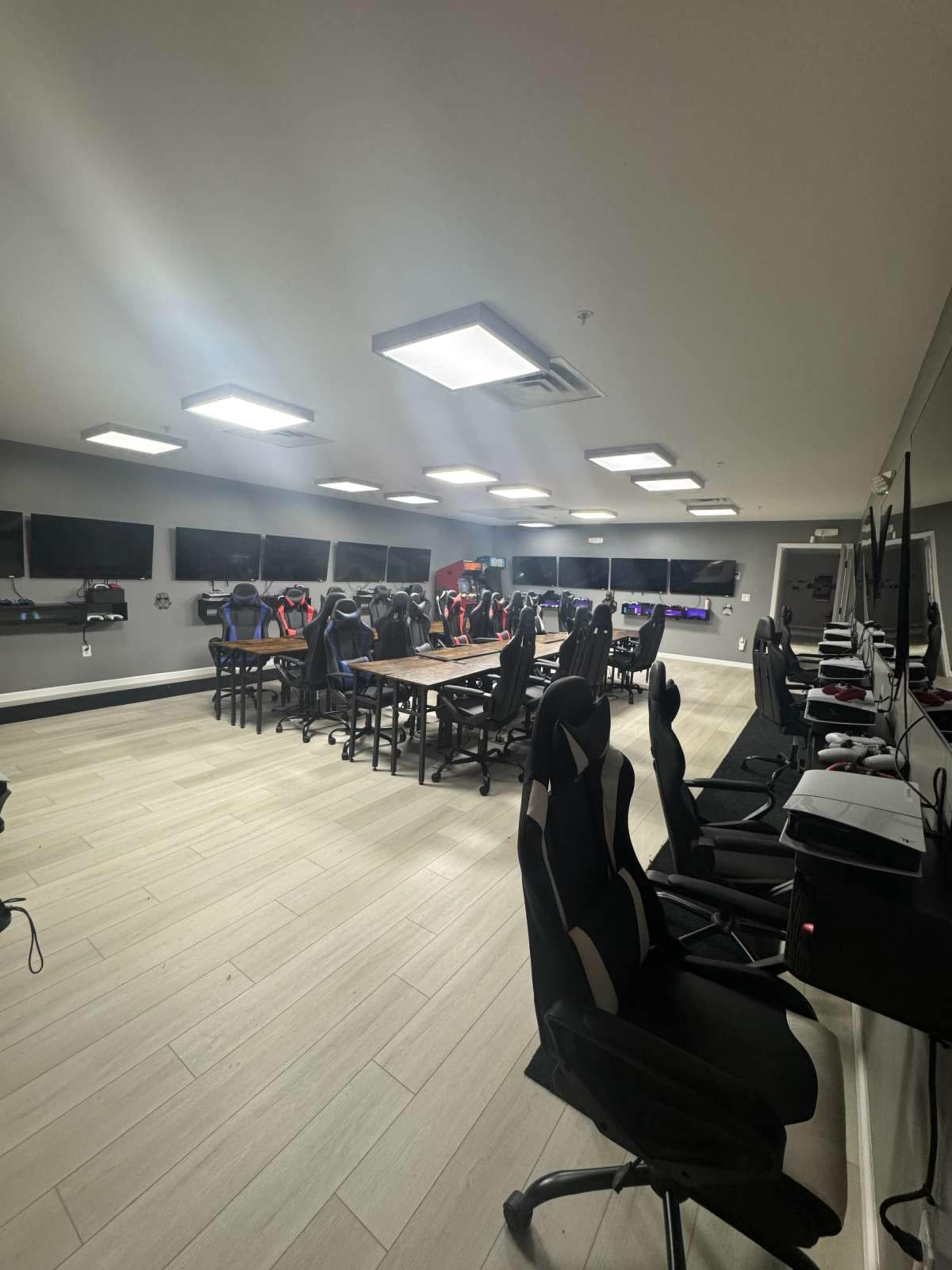 The image shows a modern gaming room with rows of gaming chairs, desks, and multiple monitors mounted on the walls.