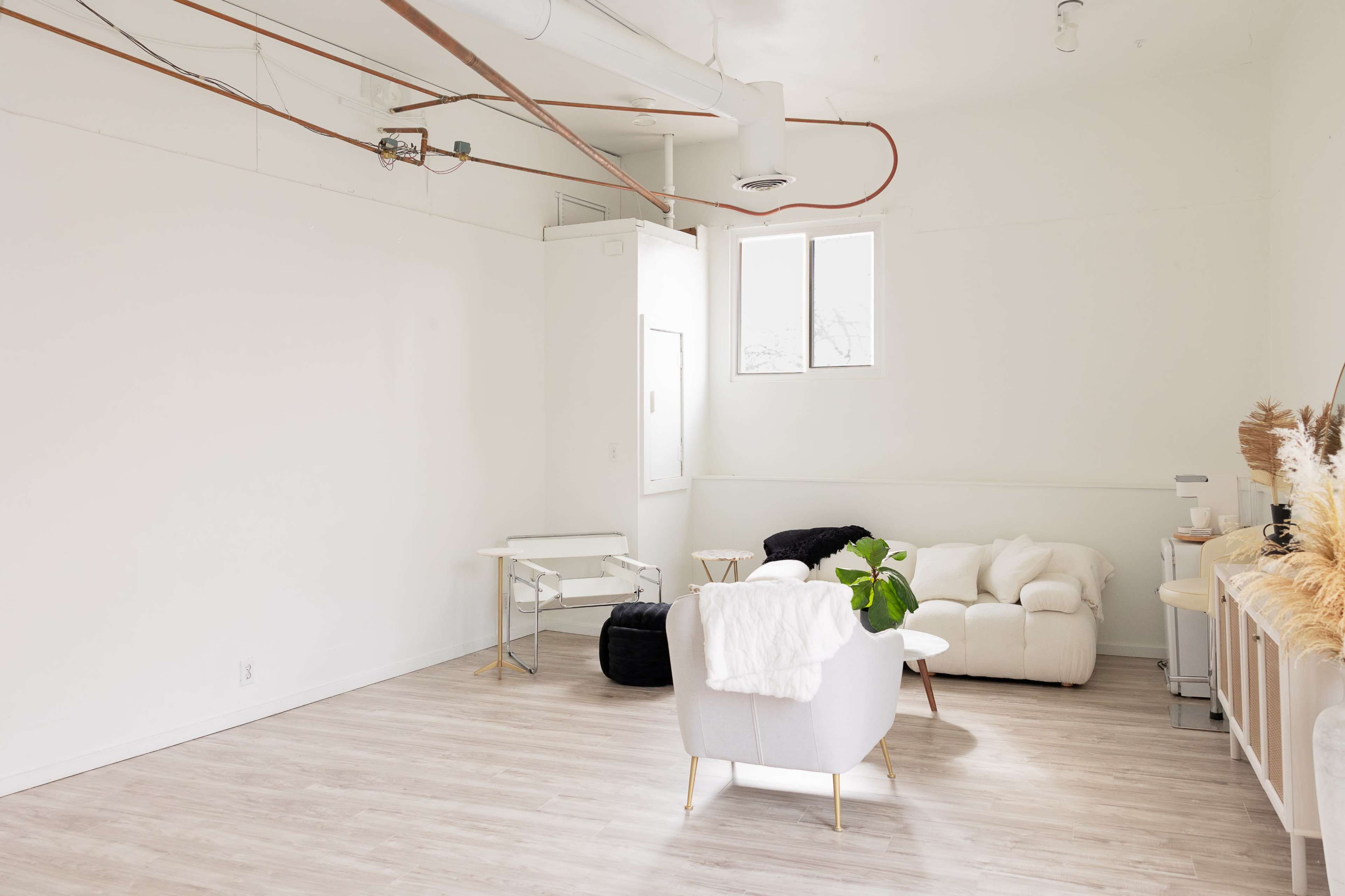 The image shows a minimalist, unfurnished room with wooden flooring, white walls, and large windows, containing a few lounge chairs and a small table.