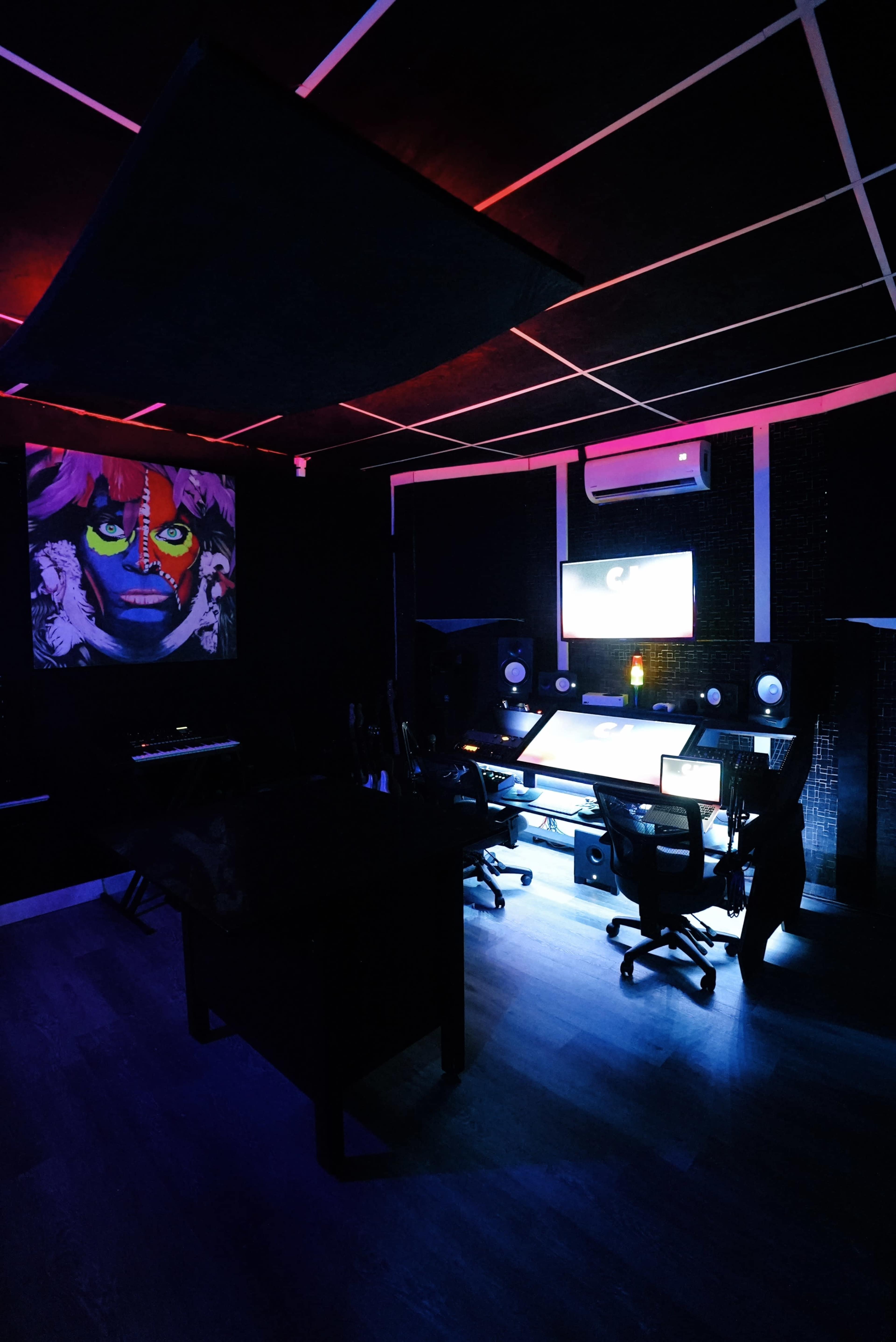 A dark music studio features colorful lighting, a large monitor, and professional audio equipment.