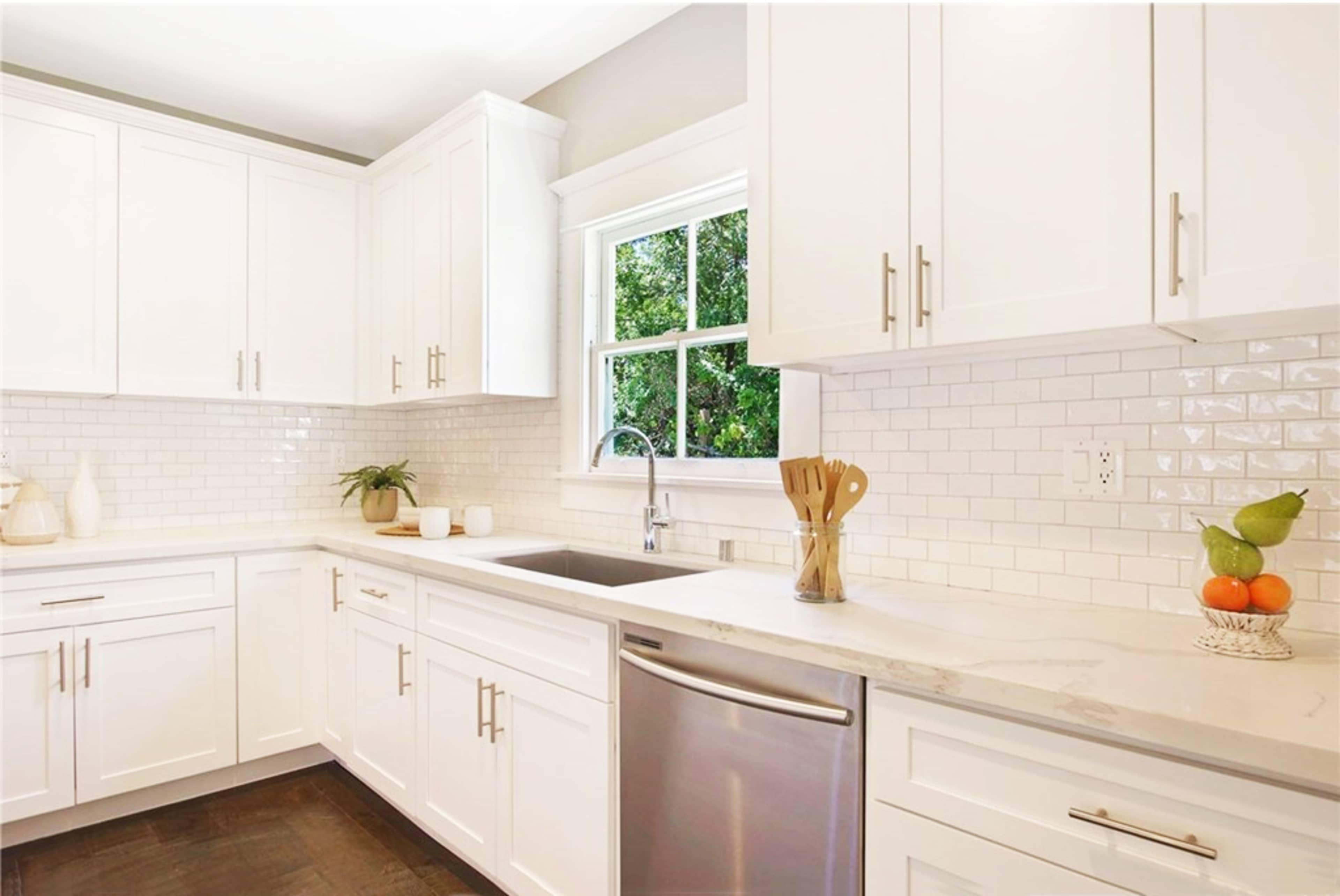 A modern kitchen features white cabinets, a stainless steel dishwasher, and a window overlooking greenery.