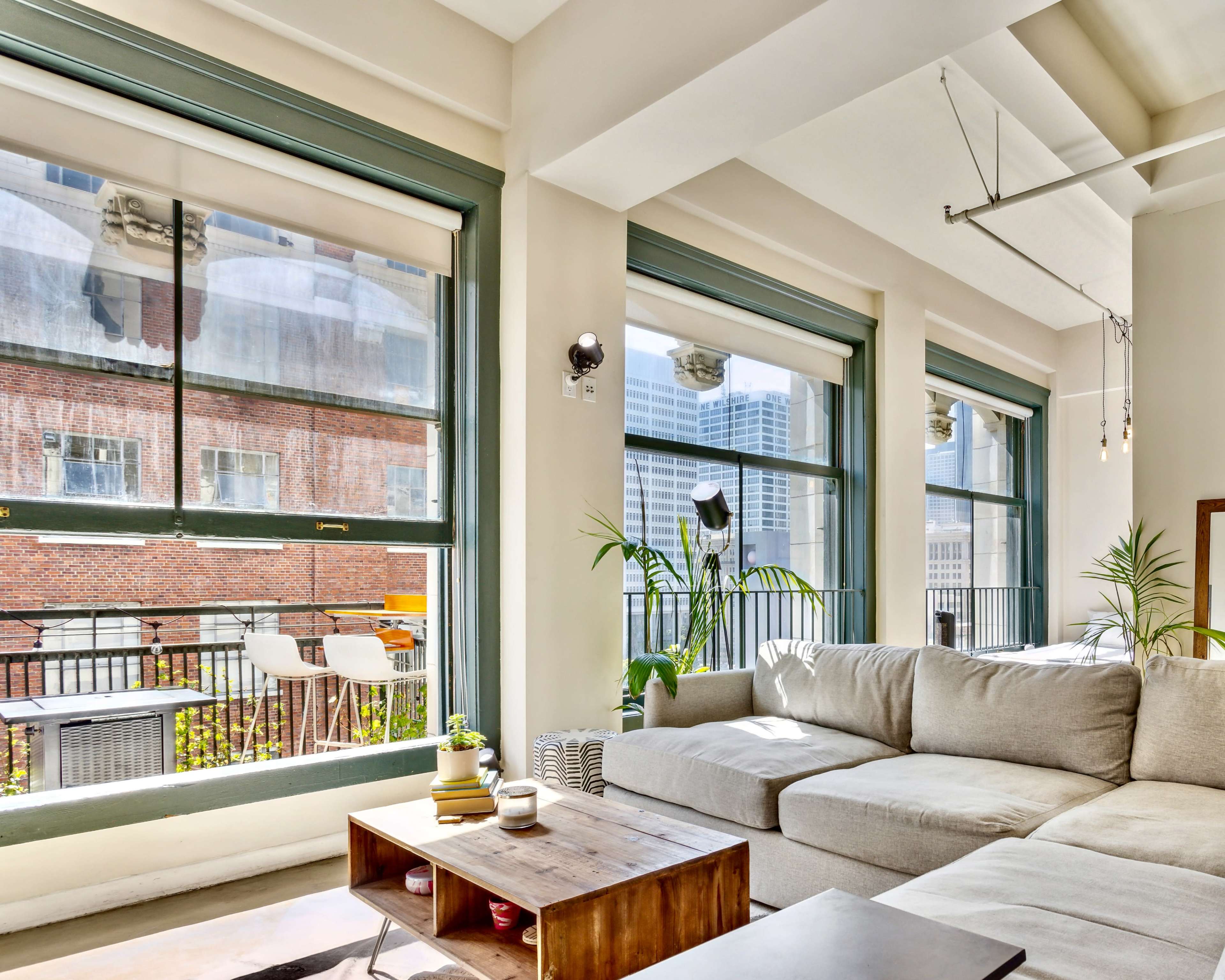 A bright, modern living room features a large sectional couch, a wooden coffee table, and expansive windows that offer views of a cityscape.