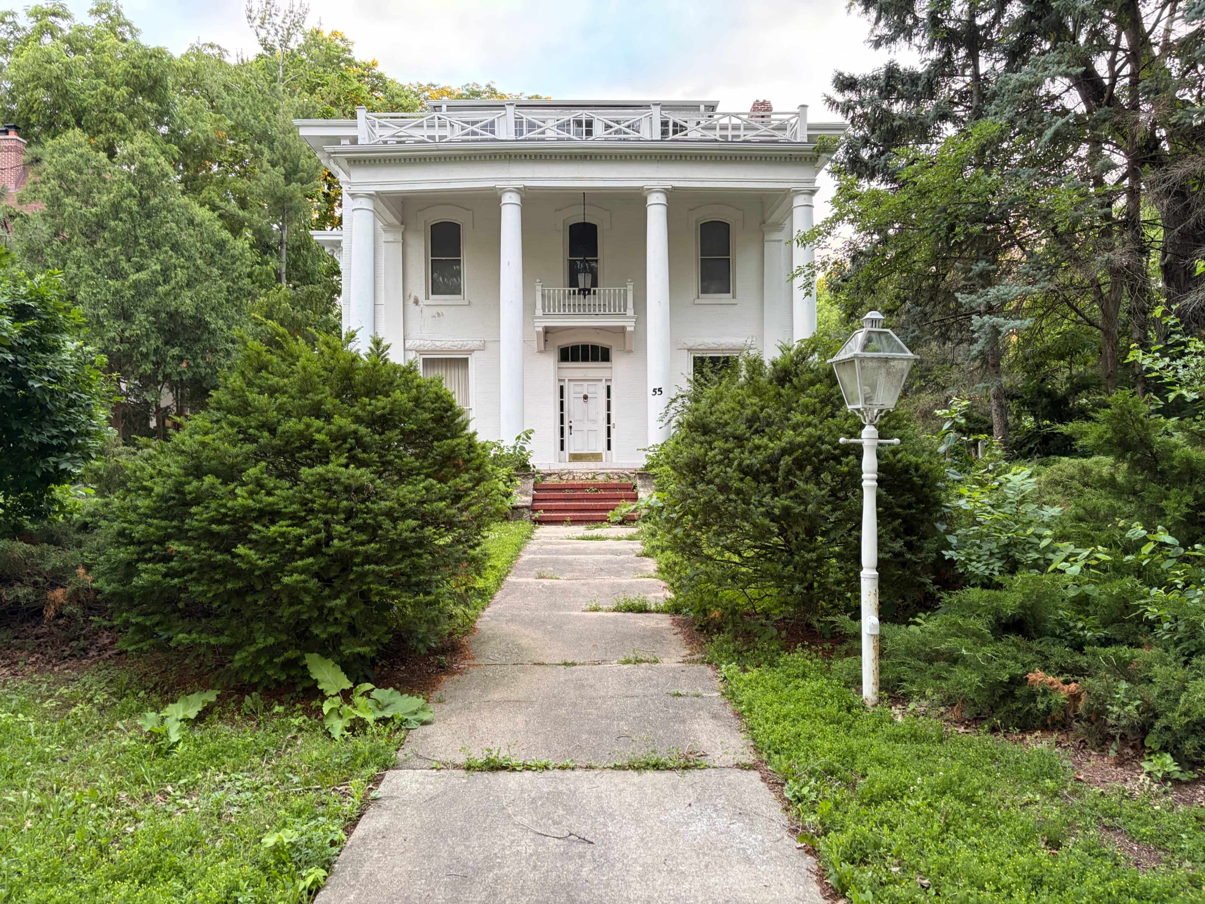 A large white house with columns stands at the end of a concrete pathway, surrounded by overgrown greenery and two neatly trimmed bushes.