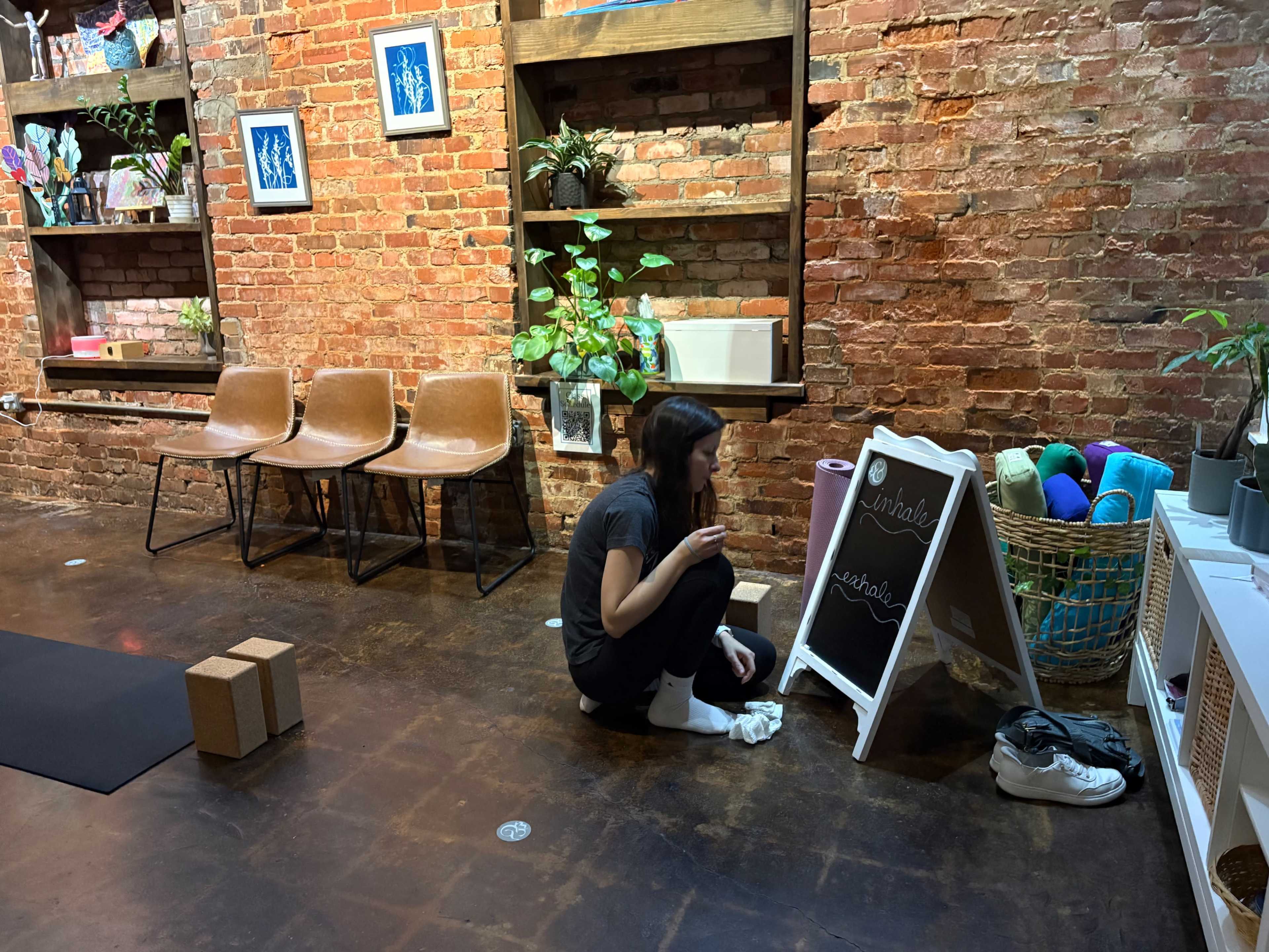 A woman kneels on the floor in a brick-walled room, writing on a chalkboard near plants and baskets of colorful materials, with chairs positioned nearby.