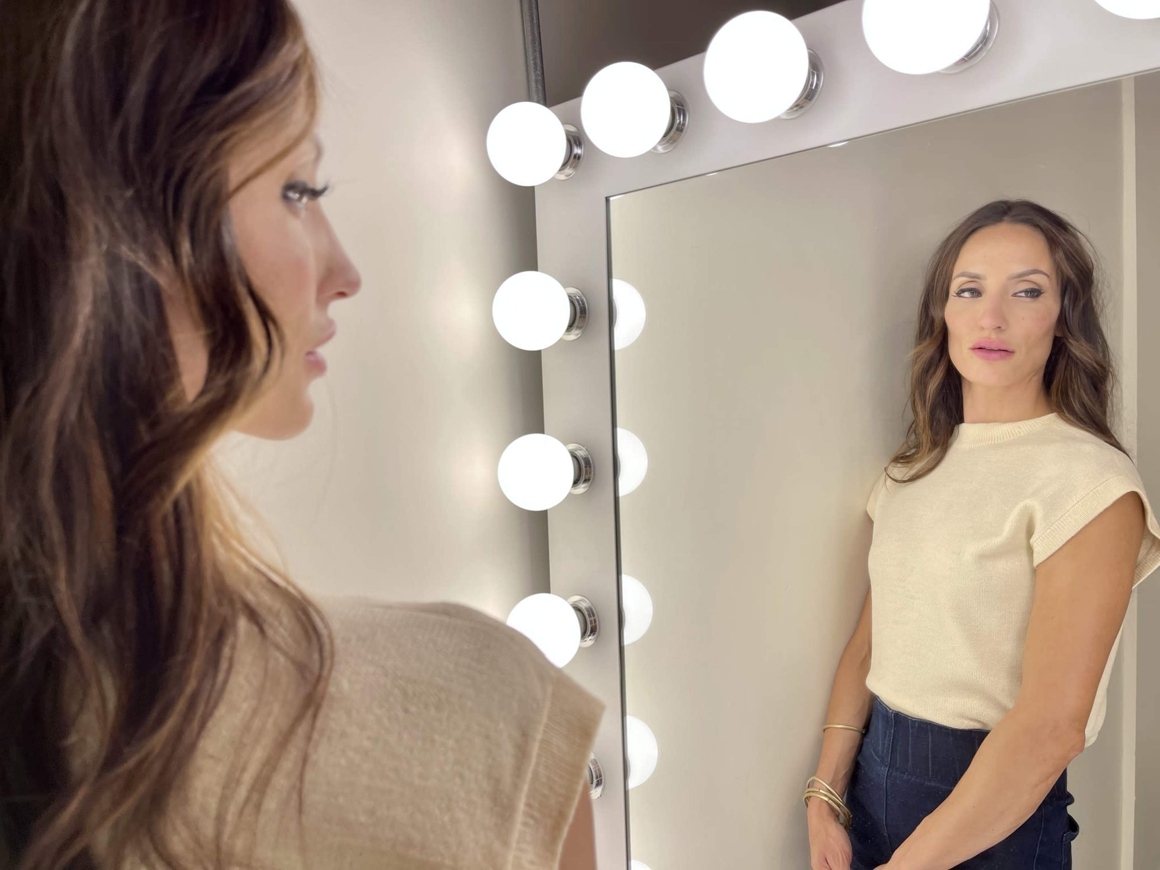 A woman stands beside a well-lit mirror, gazing at her reflection.