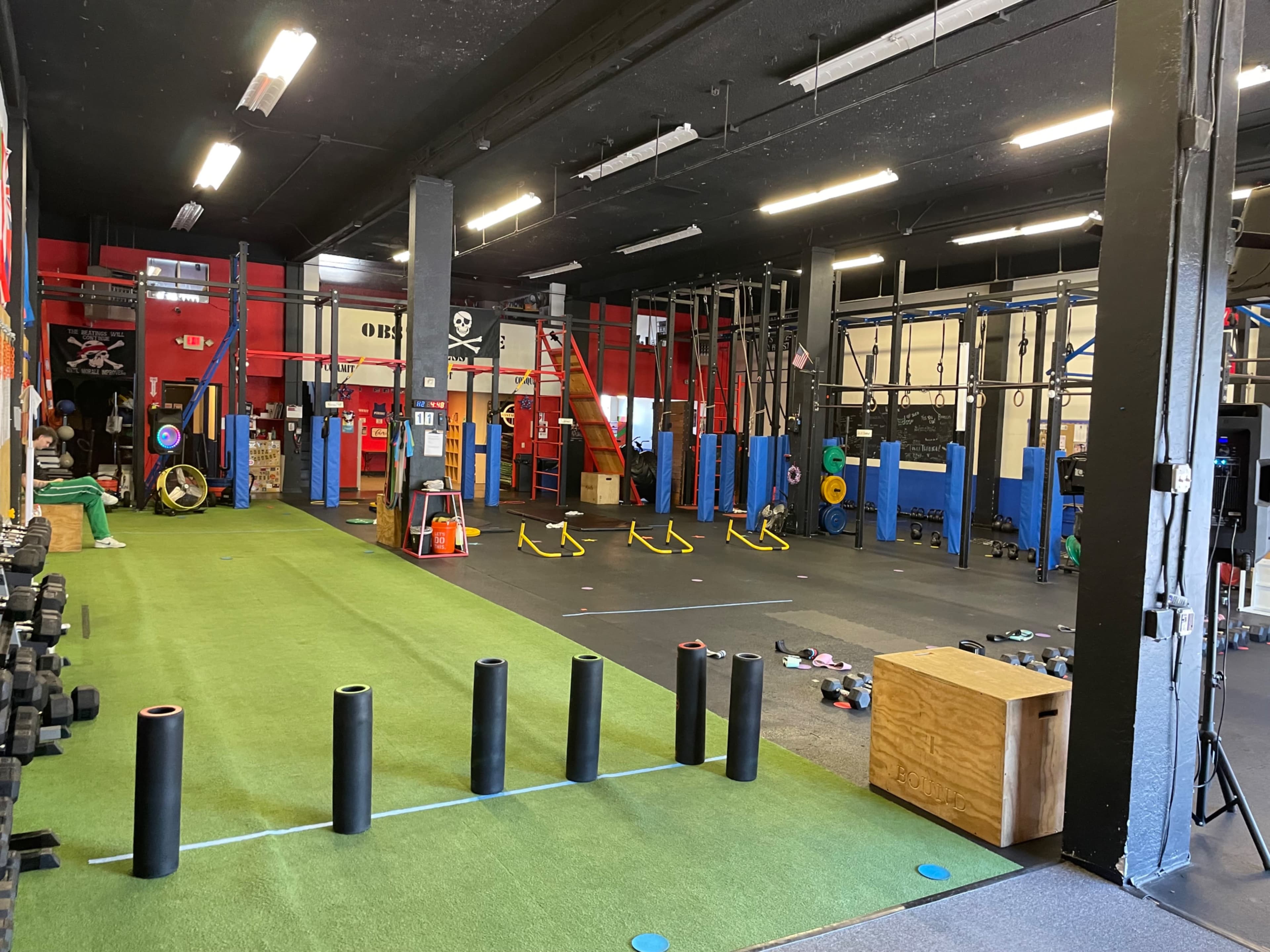 The image shows a well-equipped gym with various fitness stations, including weights, pull-up bars, and exercise mats, set up on a green turf floor.