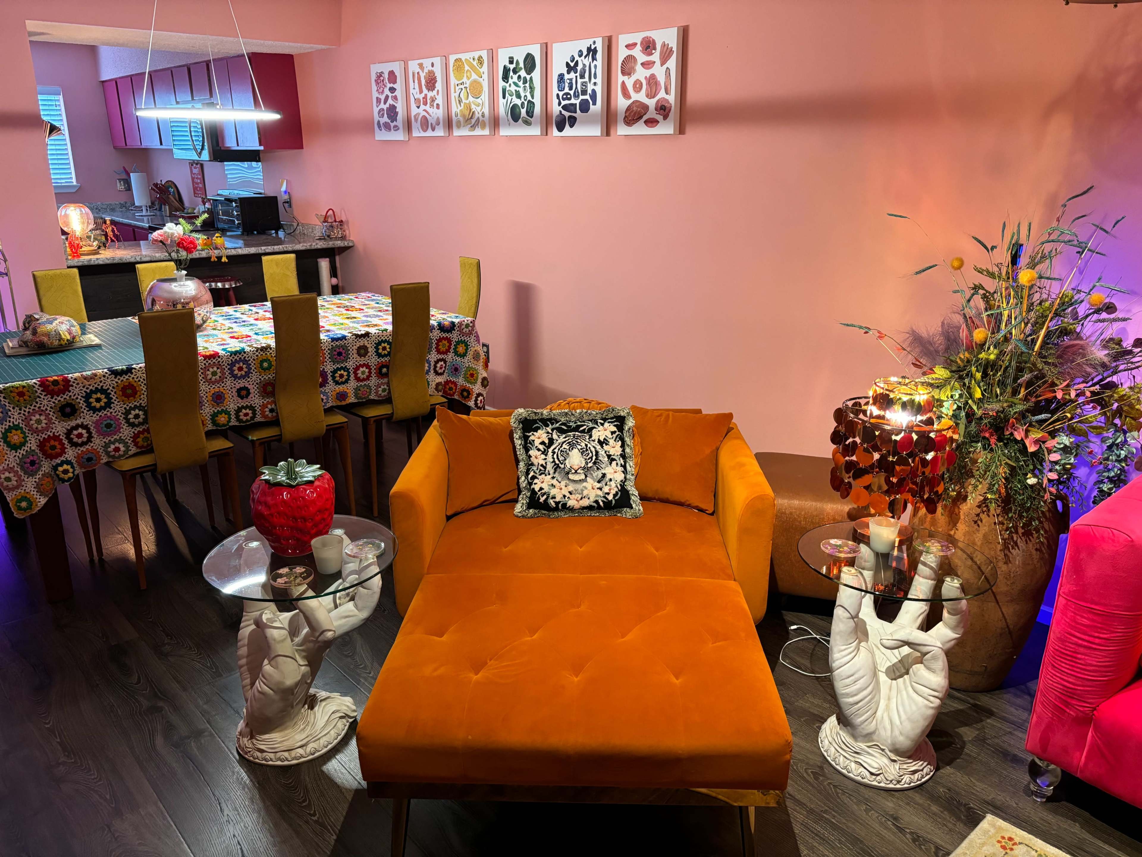 The image shows a living space with an orange chaise lounge, two side tables with decorative items, and a dining area featuring a colorful tablecloth and artwork on the pink walls.