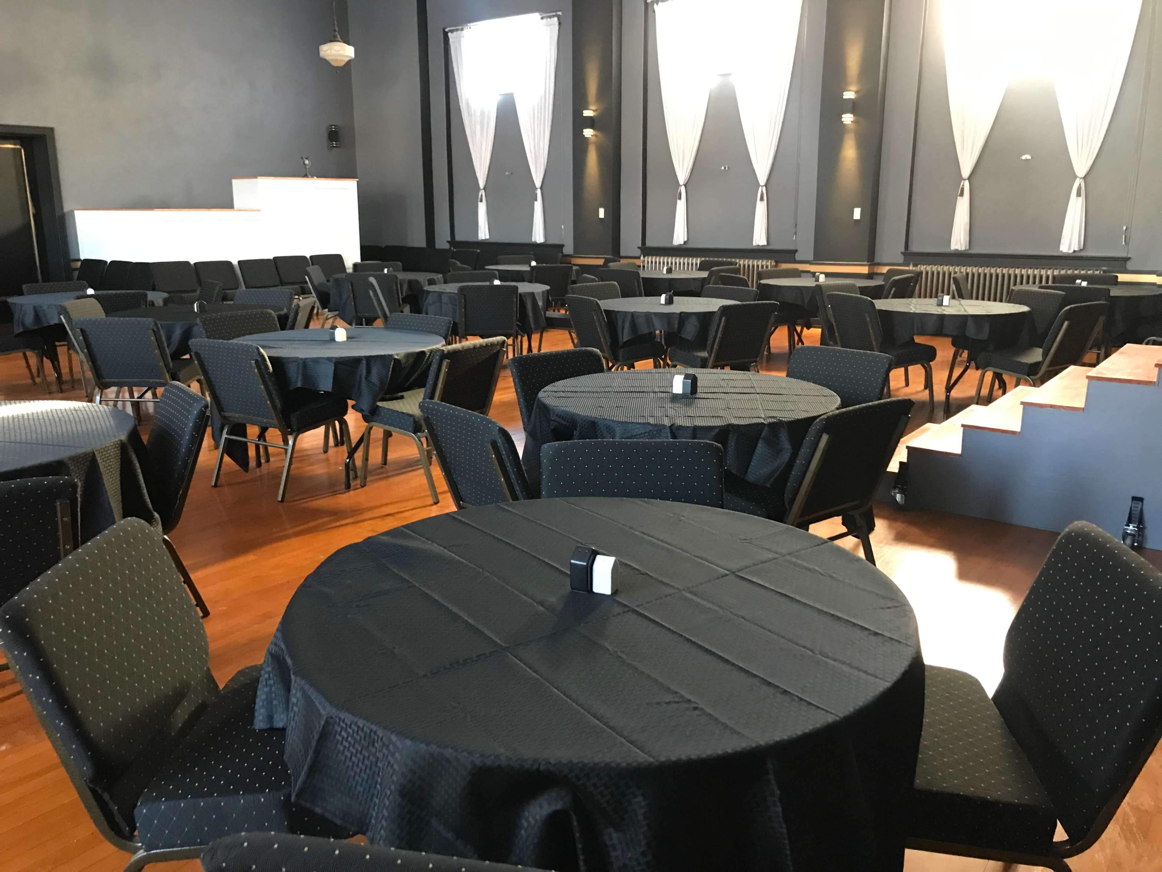 The image shows a spacious event hall set with round black tables surrounded by chairs, all arranged neatly on a polished wooden floor.