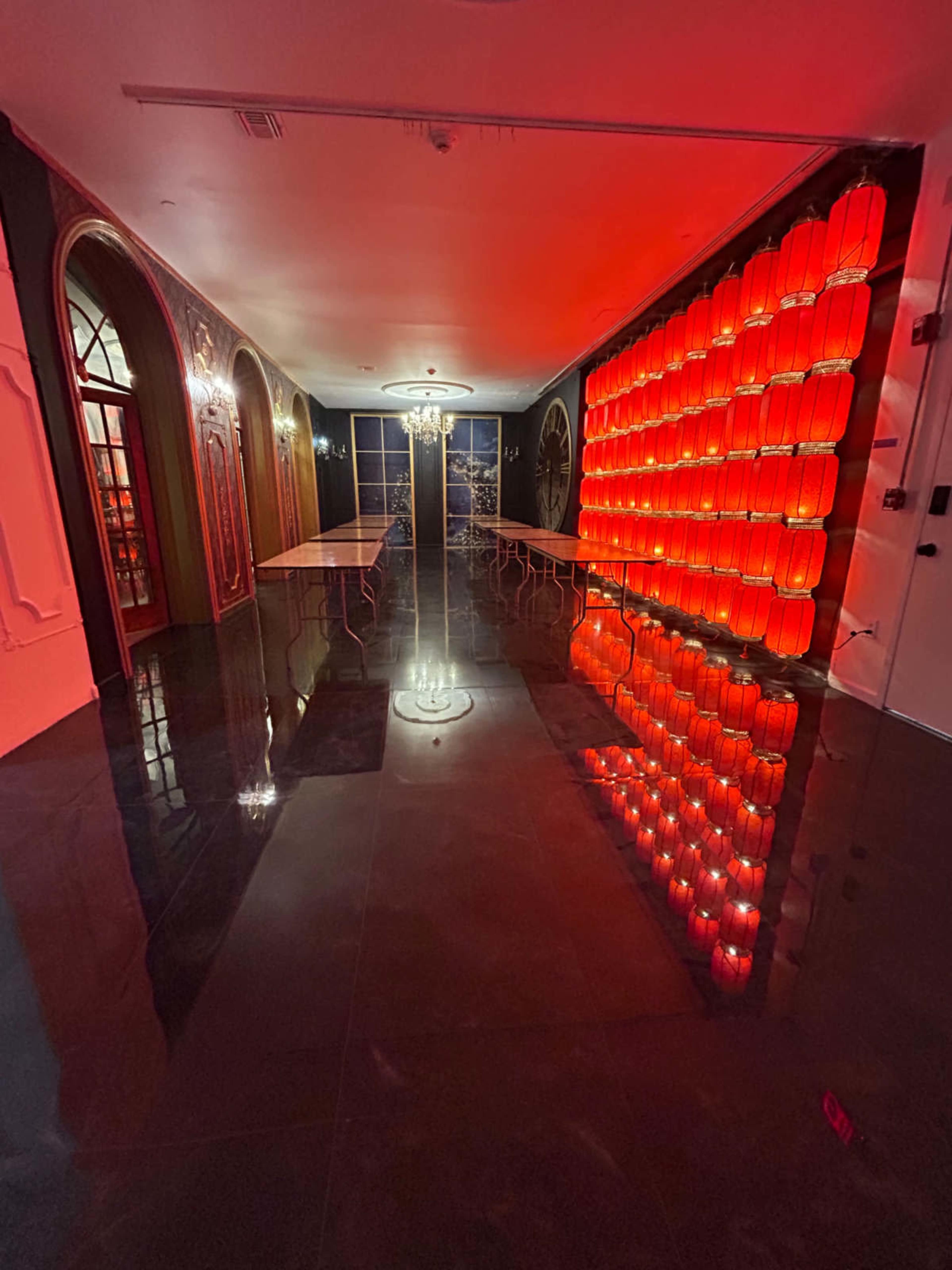 The image shows a modern indoor space with reflective floors, illuminated by red lighting, featuring a row of glowing red wall fixtures and a central chandelier.