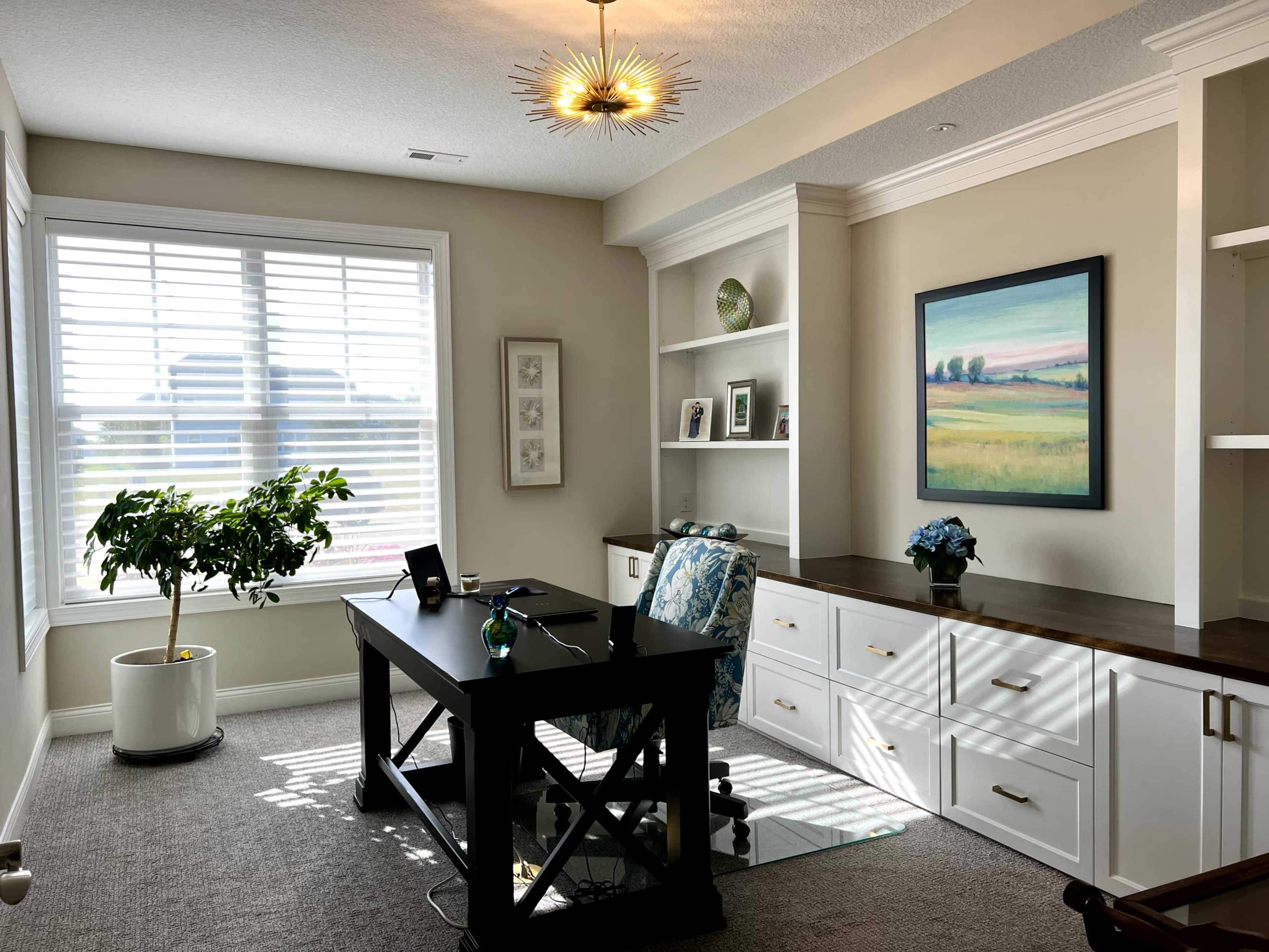 A well-lit home office features a black desk with a chair, shelves with decorative items, and a large window with white blinds.