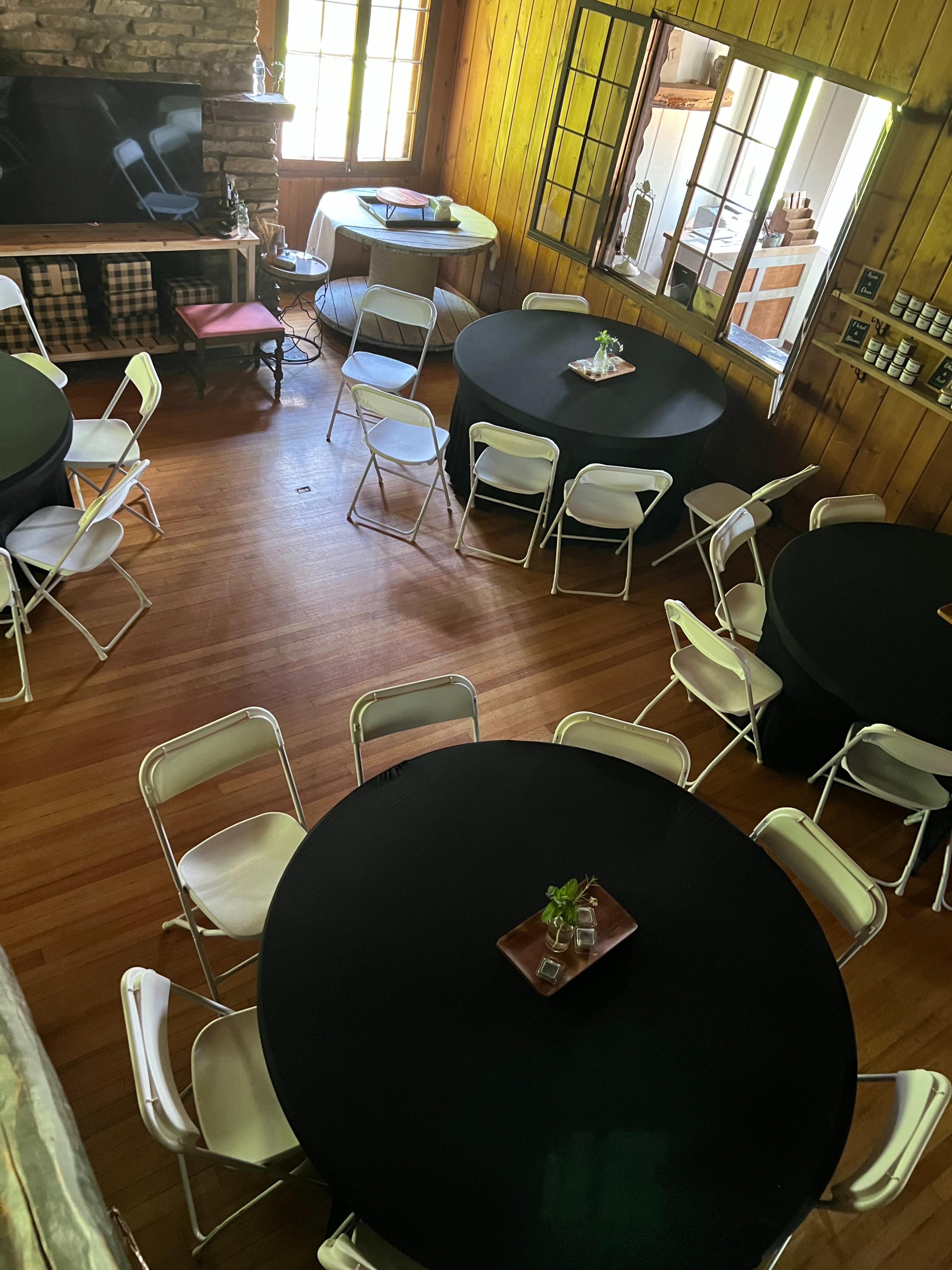The image shows a room with several round black tables surrounded by white folding chairs, with a stone fireplace and large windows.