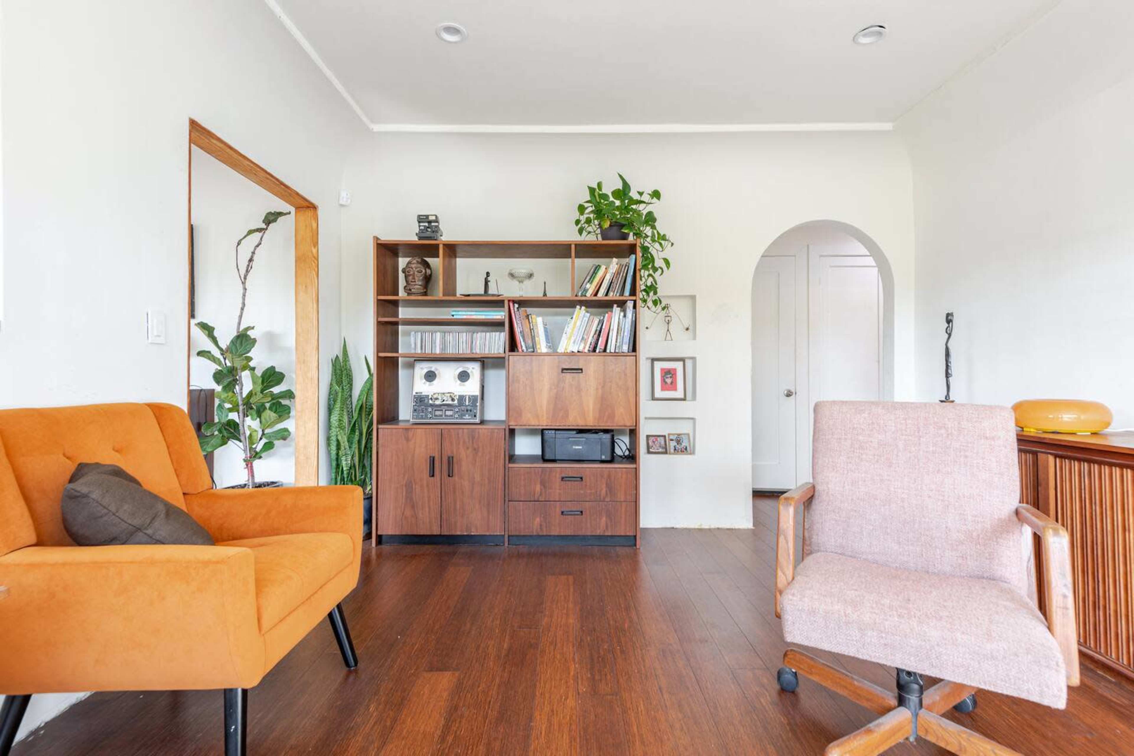 The room features two chairs, a wooden bookshelf filled with books and decorations, and plants against a light-colored wall.