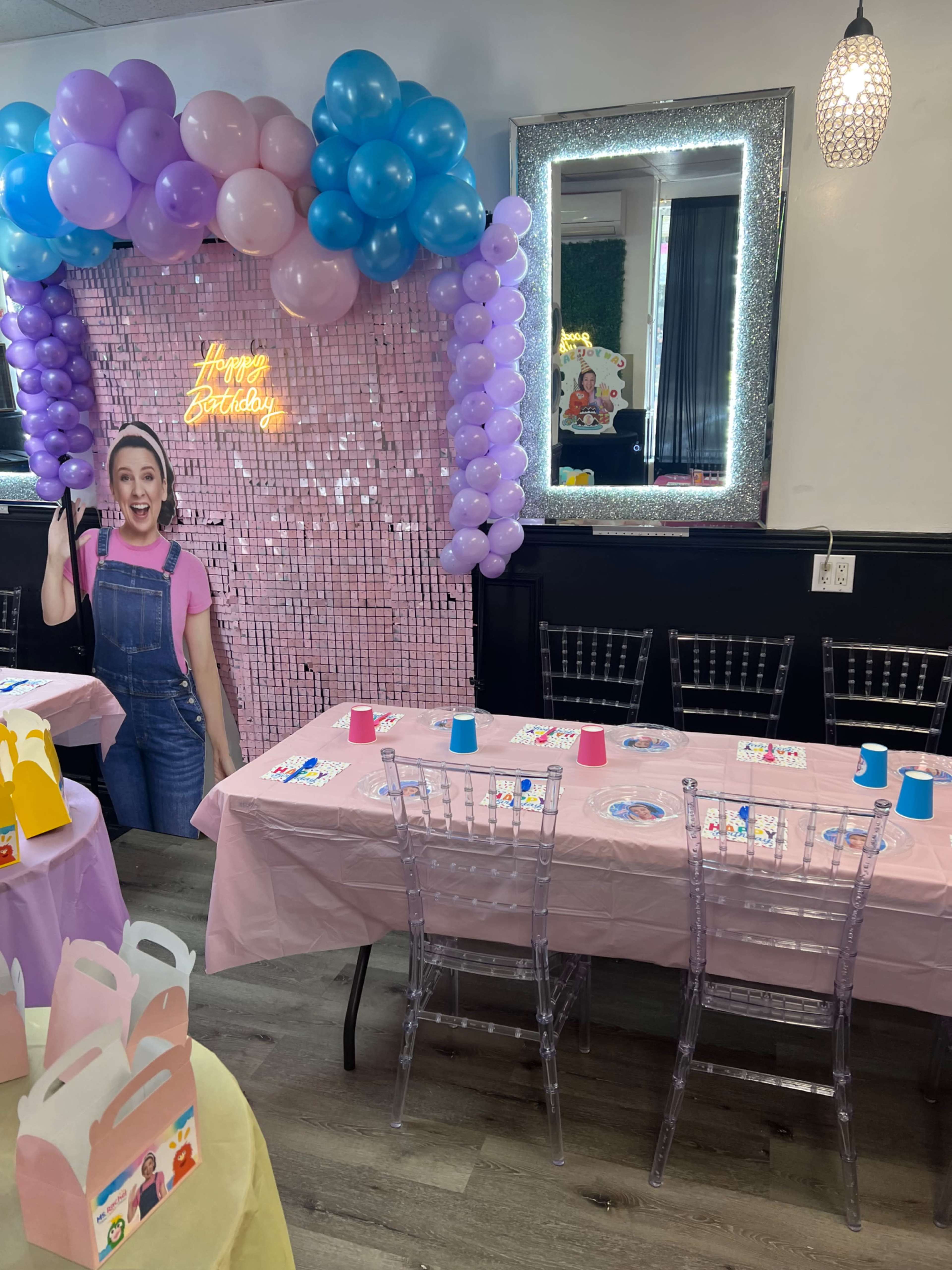 A birthday party setup features a table adorned with pink tablecloths, colorful cups, and a backdrop of balloons, while a person smiles in front of a neon "Happy Birthday" sign.