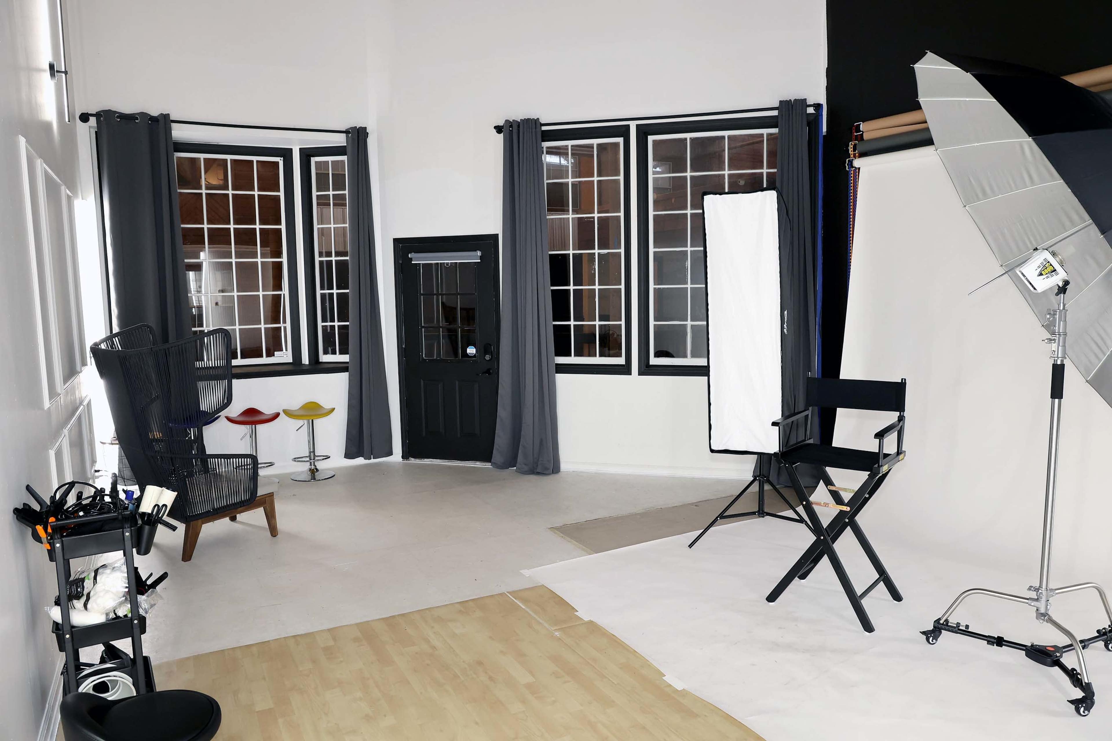 The image shows a photography studio featuring a black chair, two colorful stools, a large softbox light, and multiple windows with gray curtains.