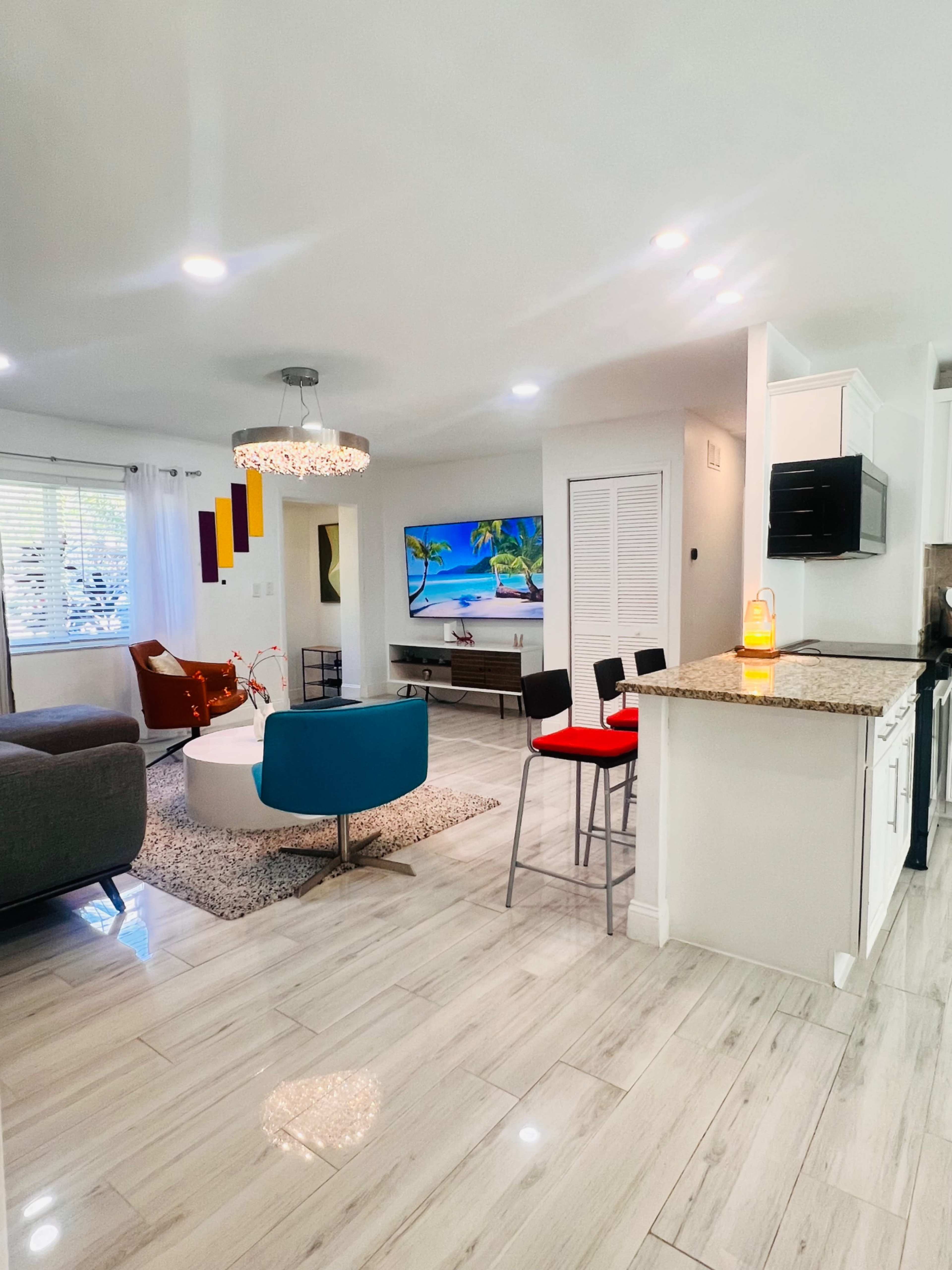 The image shows a modern living space featuring a living area with a television on the wall, a kitchen with bar stools, and a decorative chandelier.