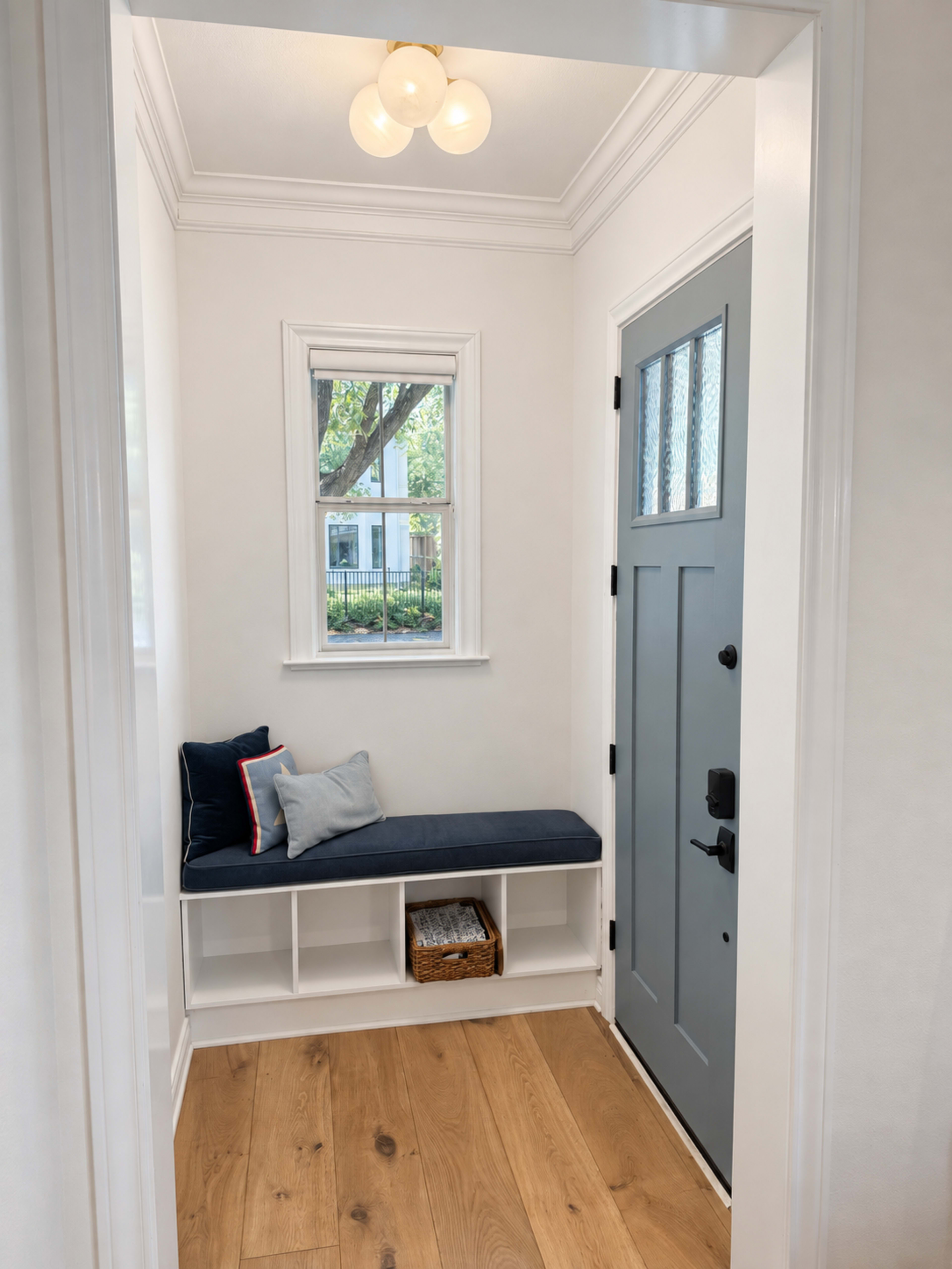 The image shows a bright entryway with a blue bench, a small decorative pillow, and a door with a window, framed by white walls and hardwood flooring.