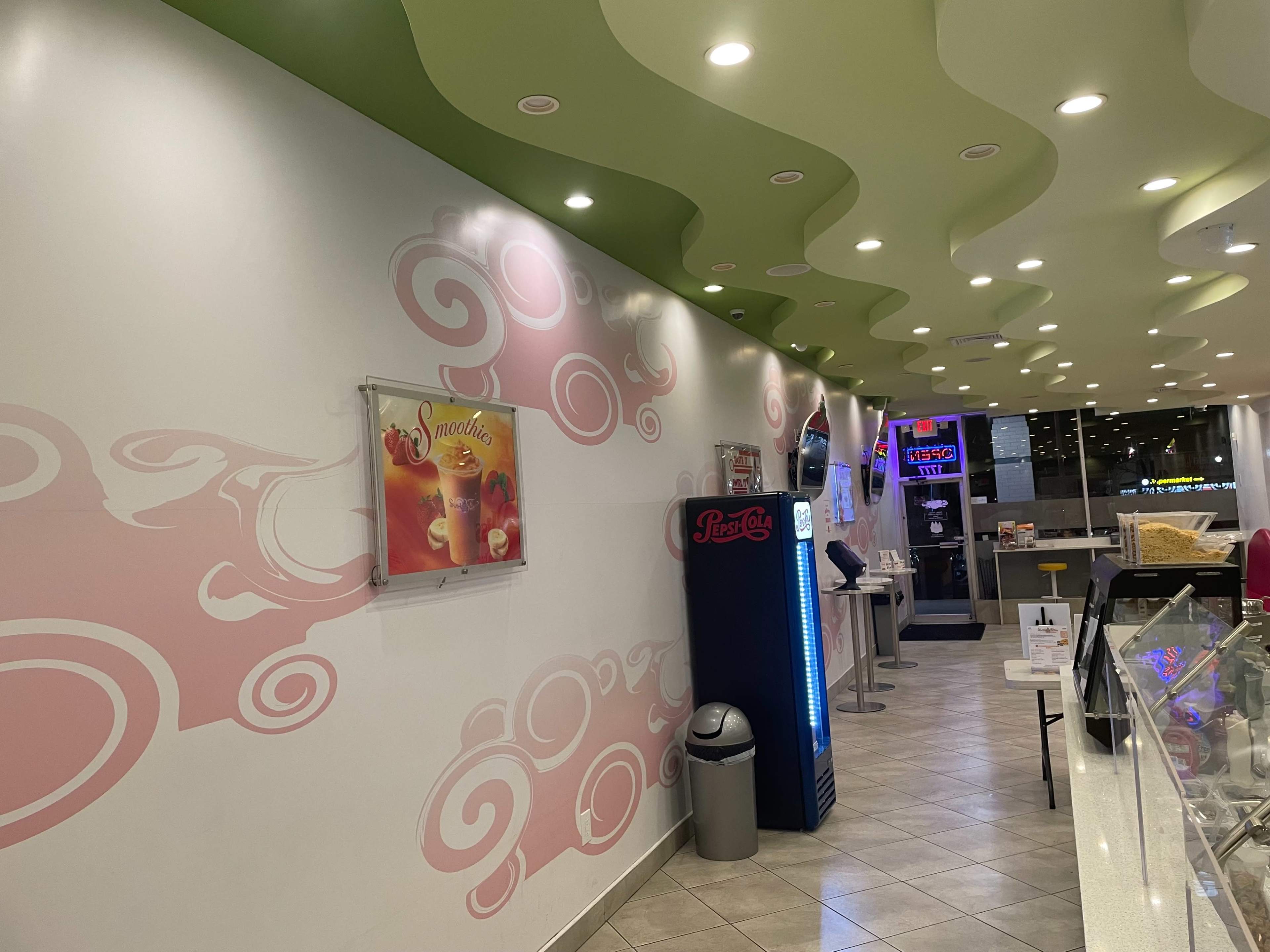 The image shows the interior of a brightly lit ice cream shop with curved green ceilings and a pink swirl motif on the walls.