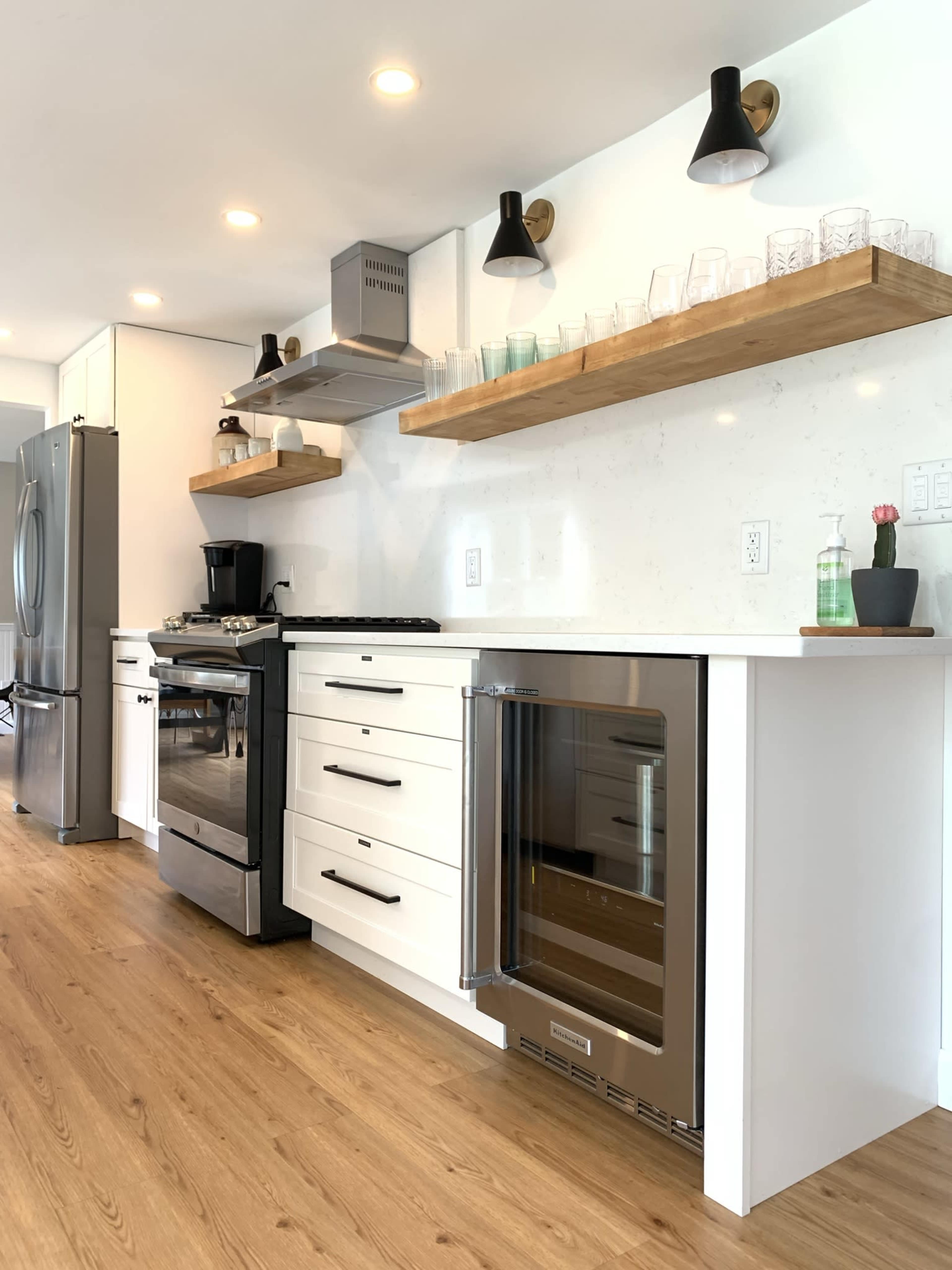 A modern kitchen with a stainless steel stove, an under-counter wine cooler, and wooden open shelving displaying glasses and a small plant.