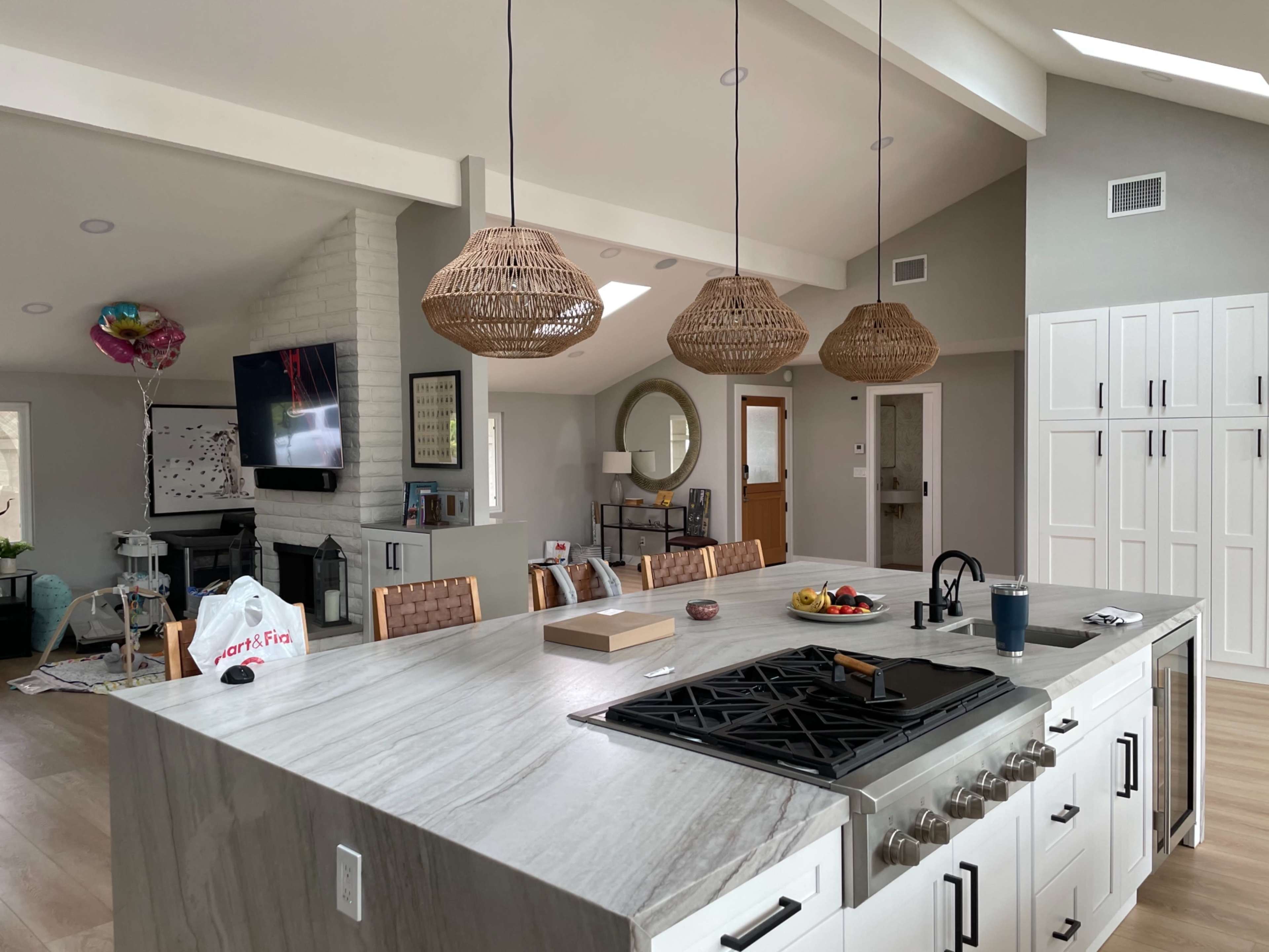 A modern kitchen features a large marble island with a gas stove, pendant lights, and an array of kitchen appliances and colorful fruit on the counter.