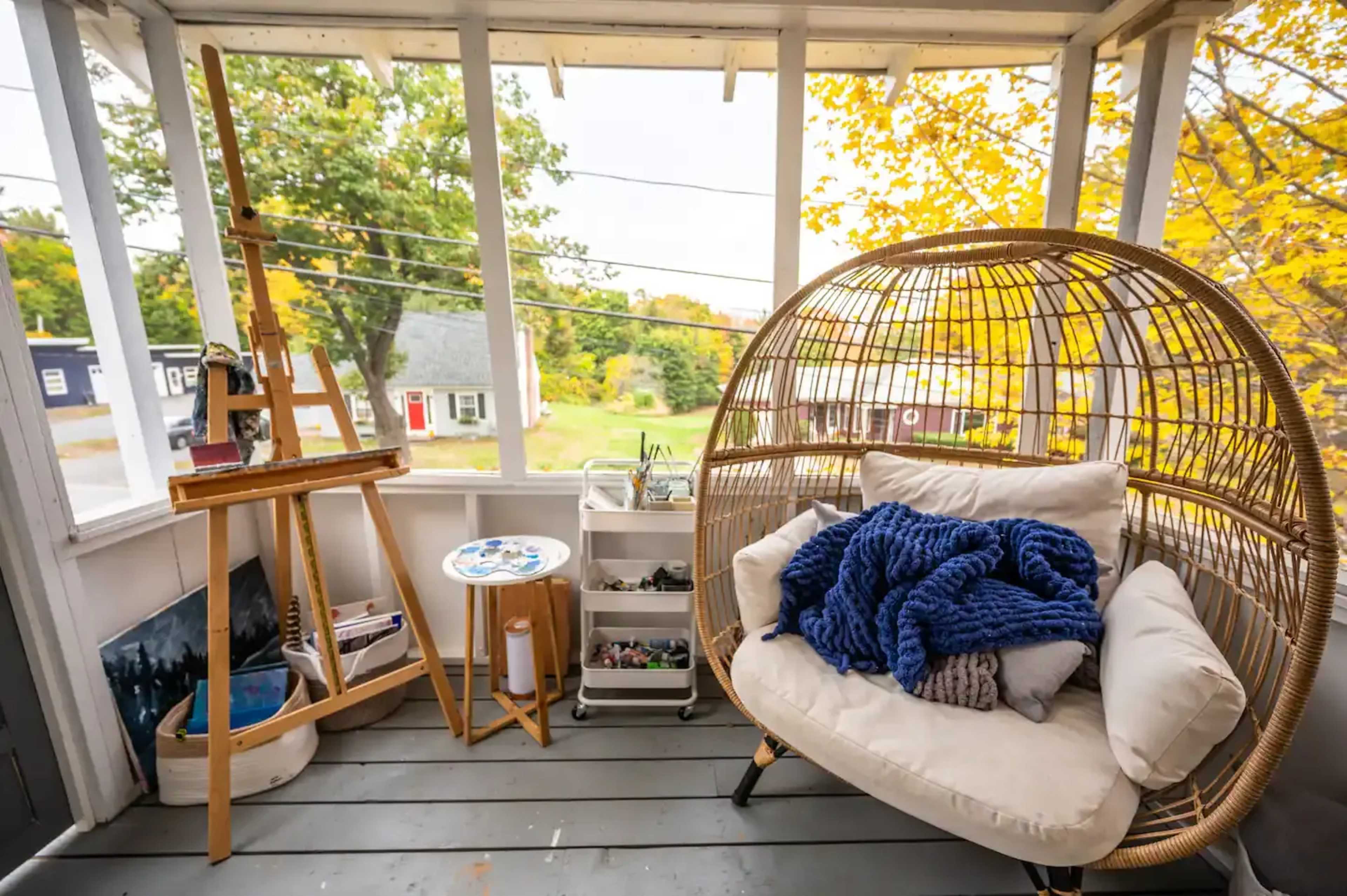 A cozy sunroom features a large wicker chair with cushions, an easel, and a table, all set against a backdrop of autumn-colored trees outside.