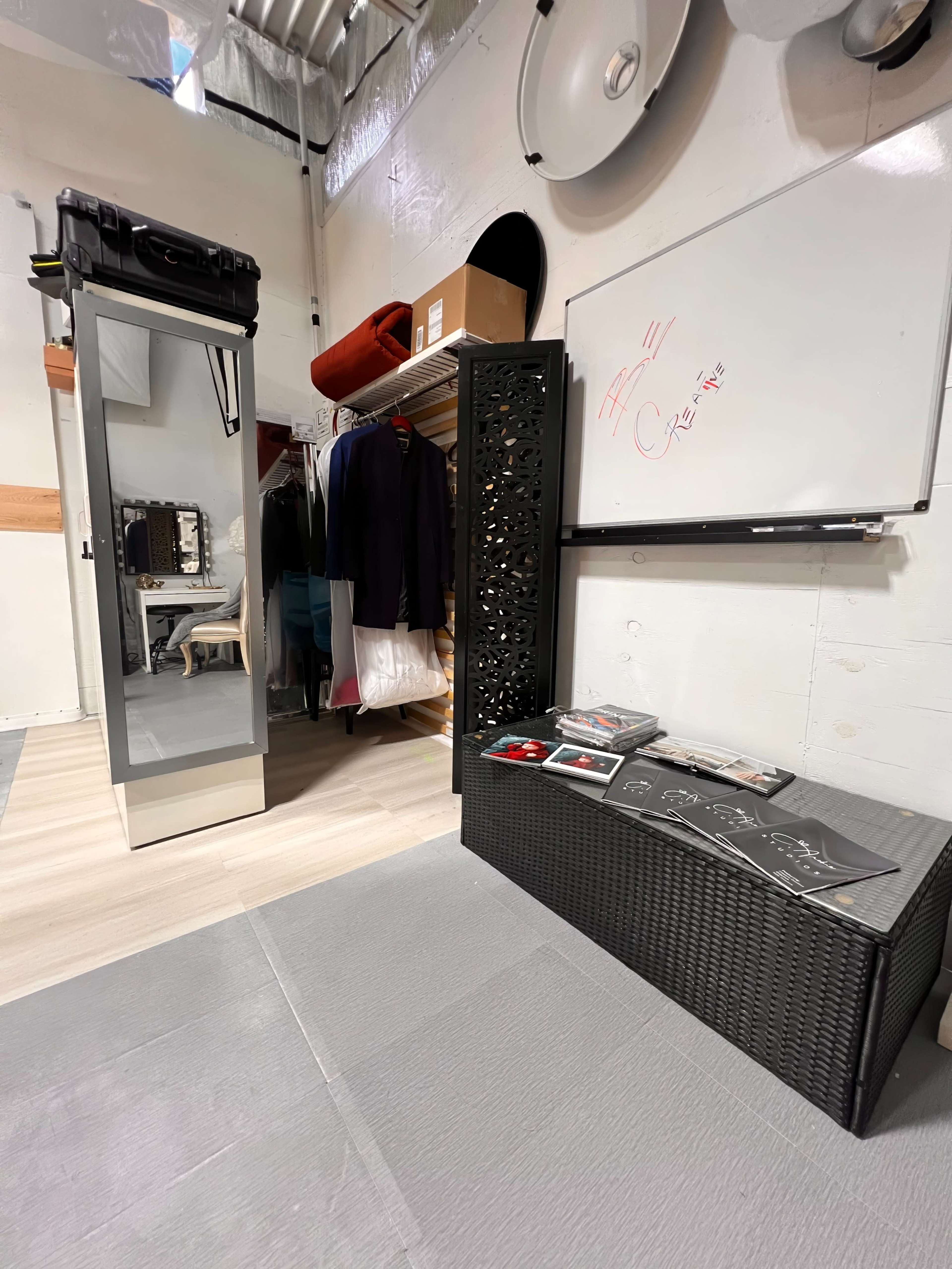 A storage area with a full-length mirror, clothing on hangers, a whiteboard, and a wicker storage box with various items on top.