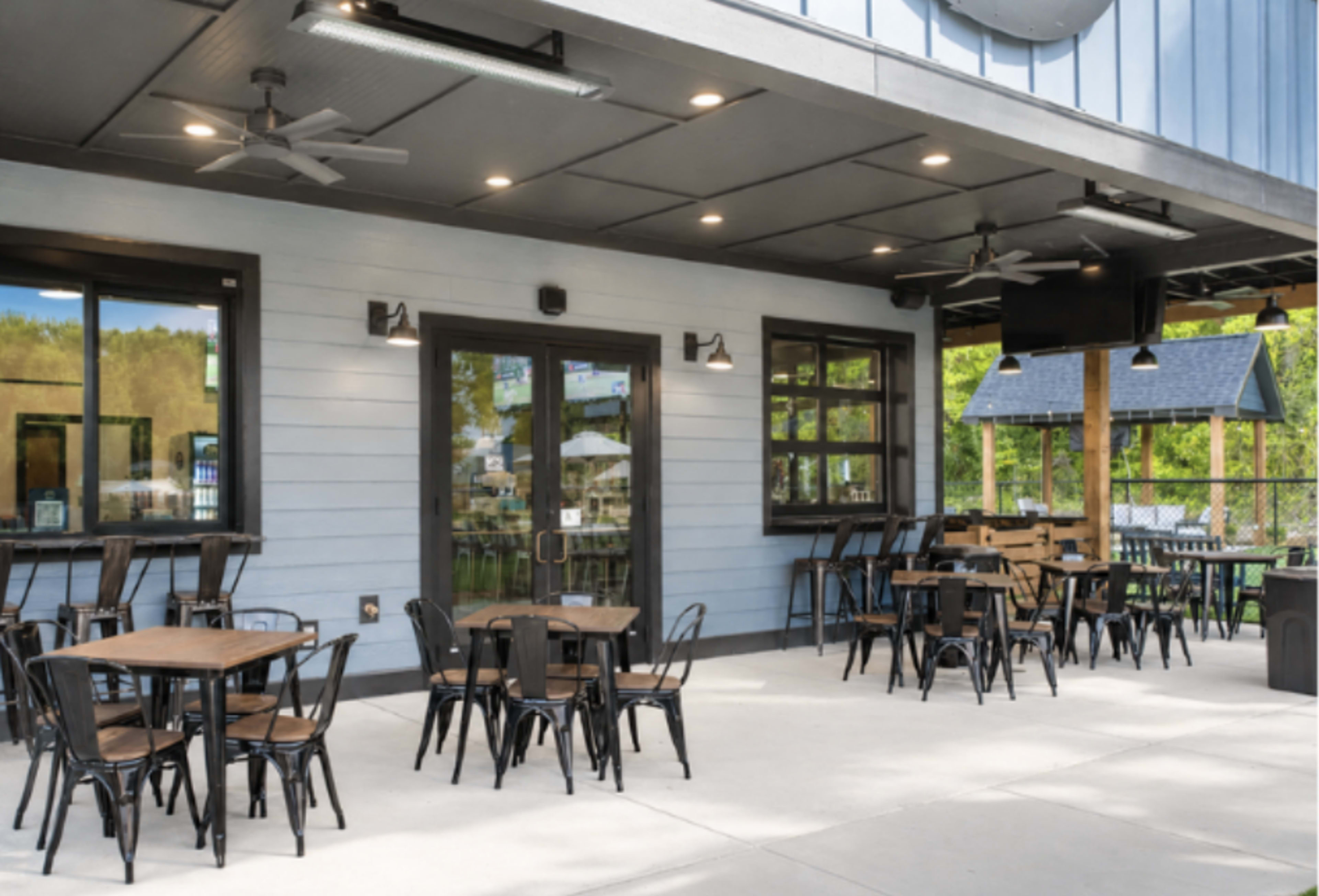 The image depicts an outdoor dining area featuring several tables and chairs arranged on a concrete patio, adjacent to a building with large windows and overhead fans.