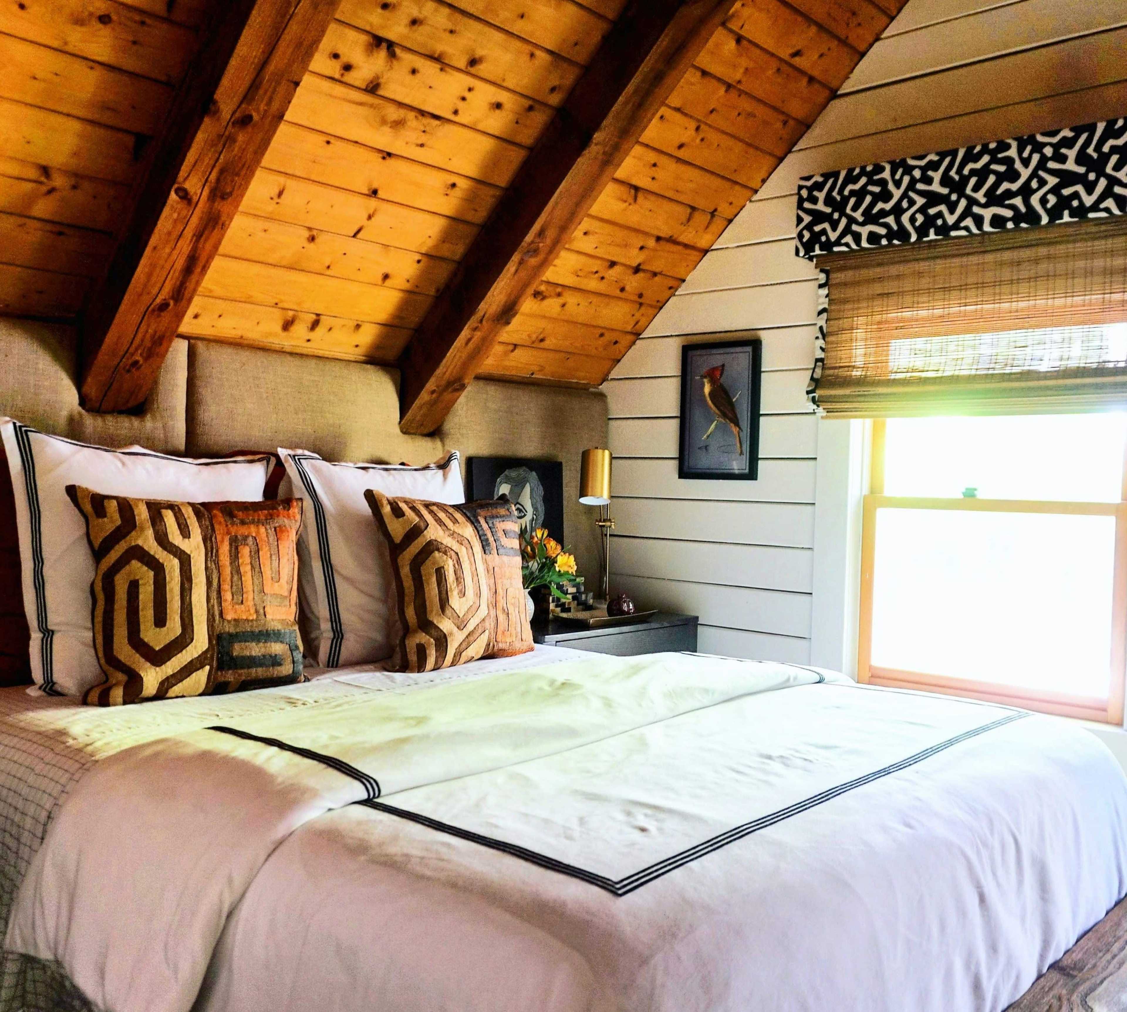 A cozy bedroom with a sloped wooden ceiling, a large bed adorned with patterned pillows, and a window allowing natural light to enter.
