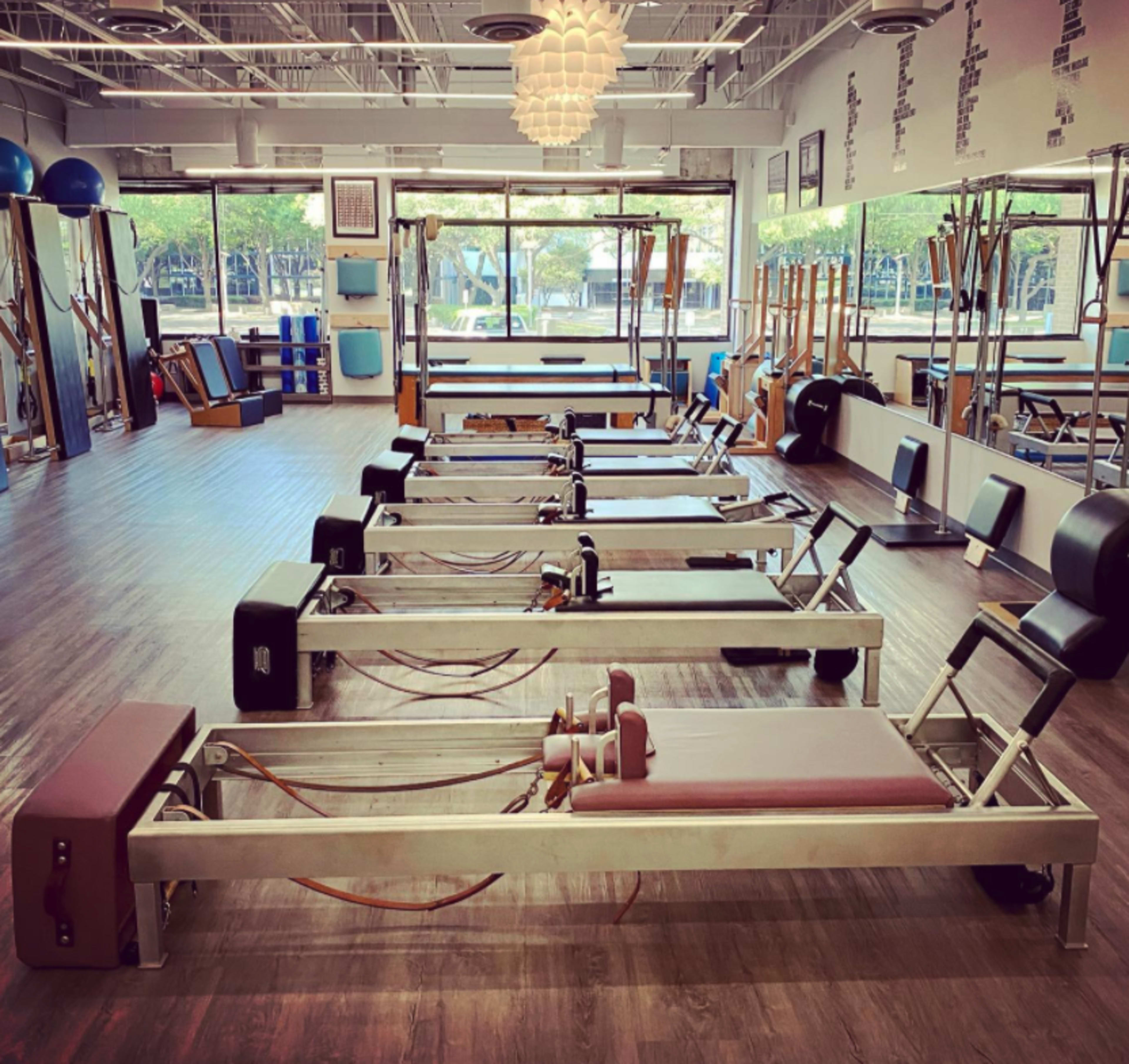 The image shows a bright, spacious pilates studio featuring several reformer machines arranged neatly along a wooden floor.