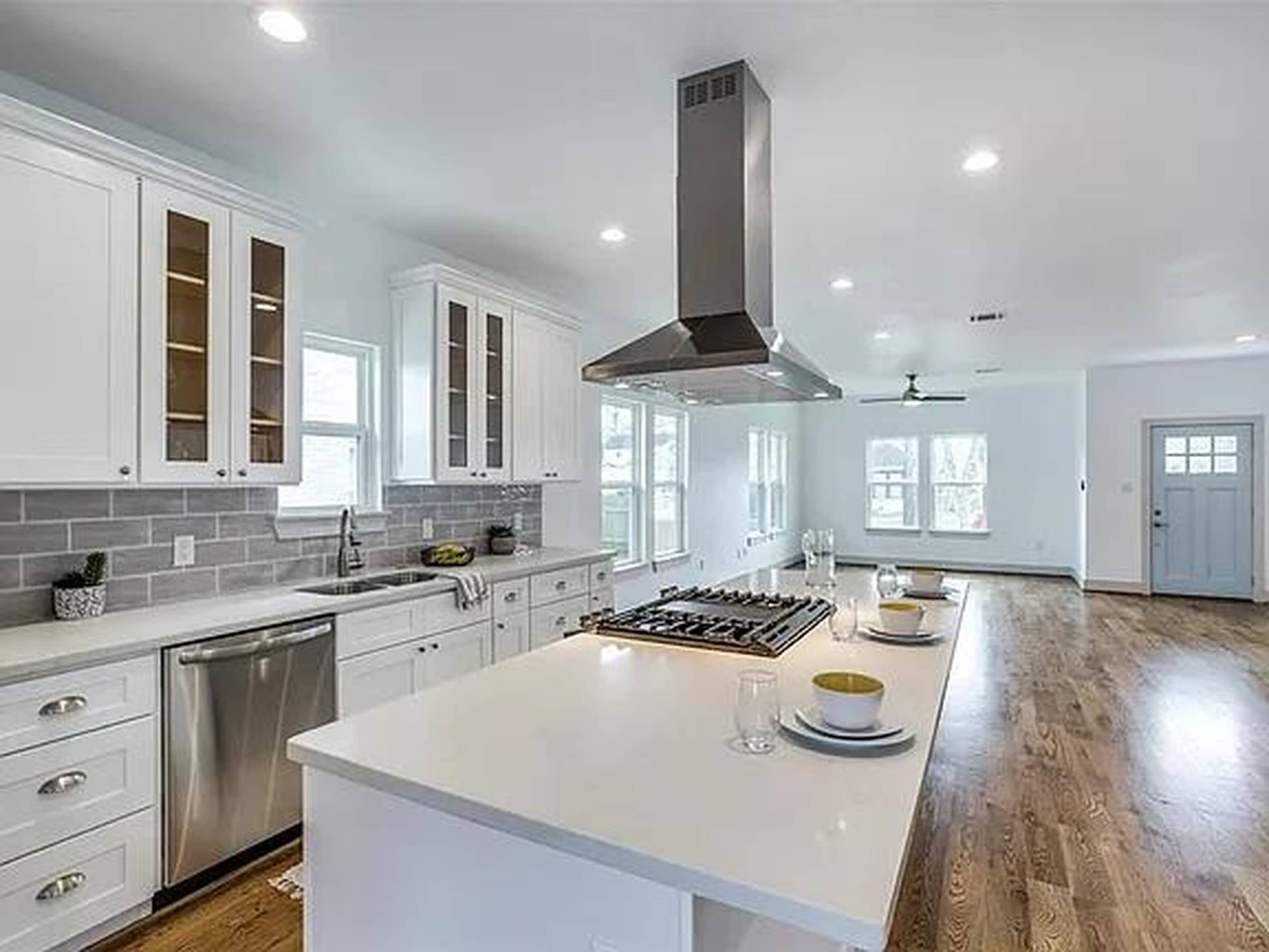 The image shows a modern kitchen with white cabinetry, a central island with a stove, stainless steel appliances, and hardwood flooring.
