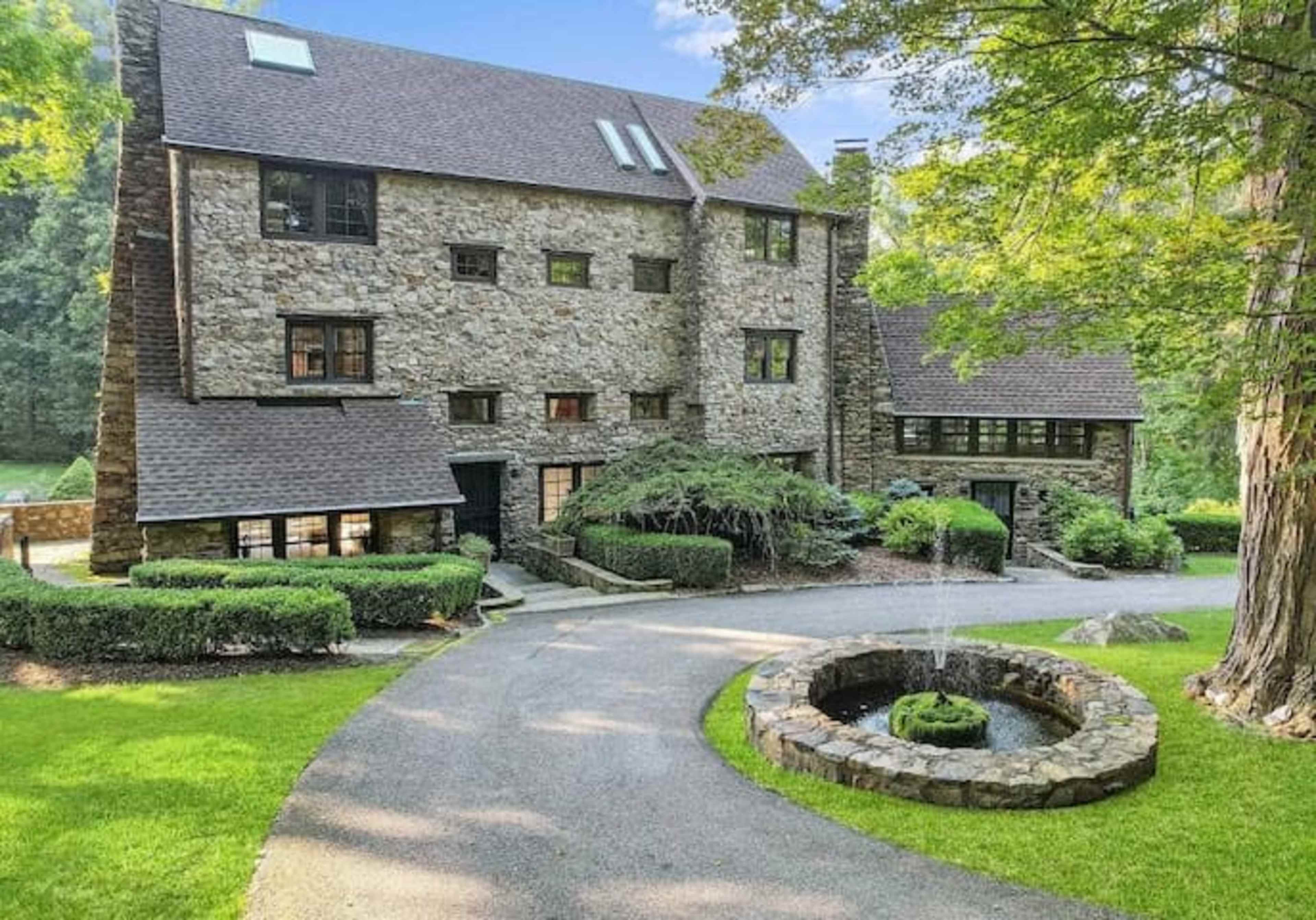 A multi-story stone house with a circular driveway and a fountain surrounded by a landscaped yard.