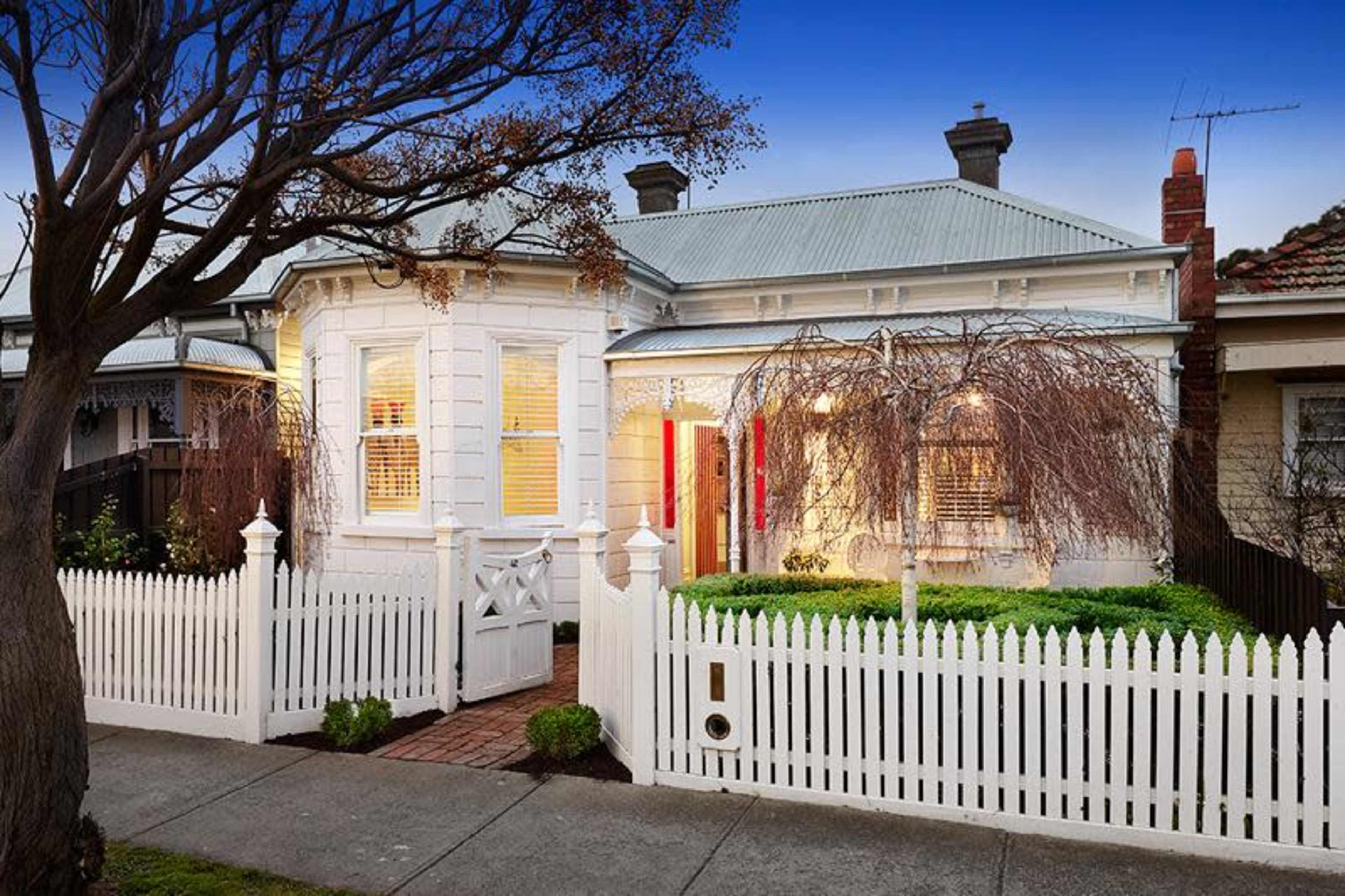 A white, single-story house with a curved facade, surrounded by a white picket fence and greenery, is shown at dusk.