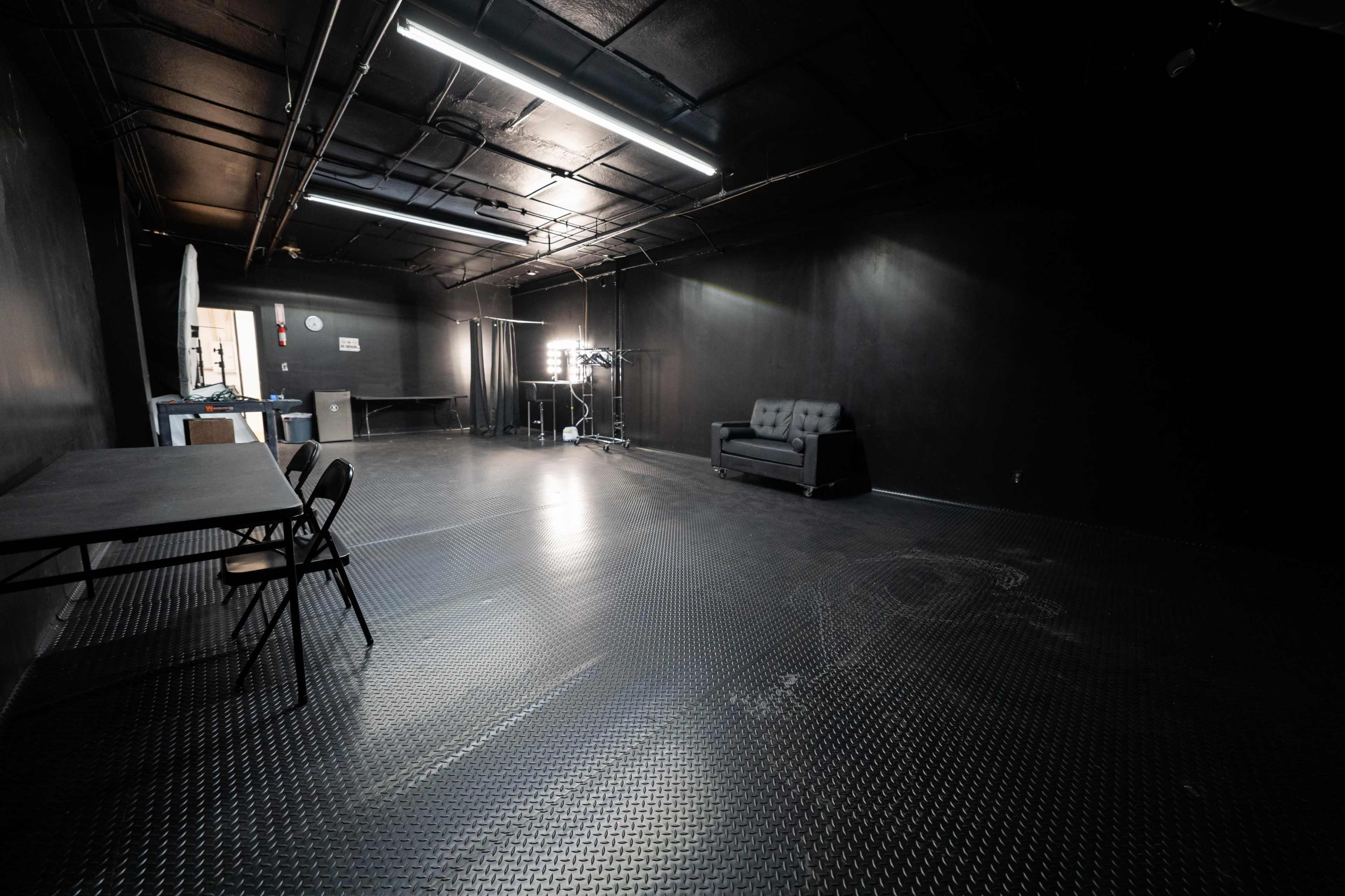 A dimly lit, empty room with black walls and a textured floor, featuring a folding table with chairs and a gray couch along the wall.
