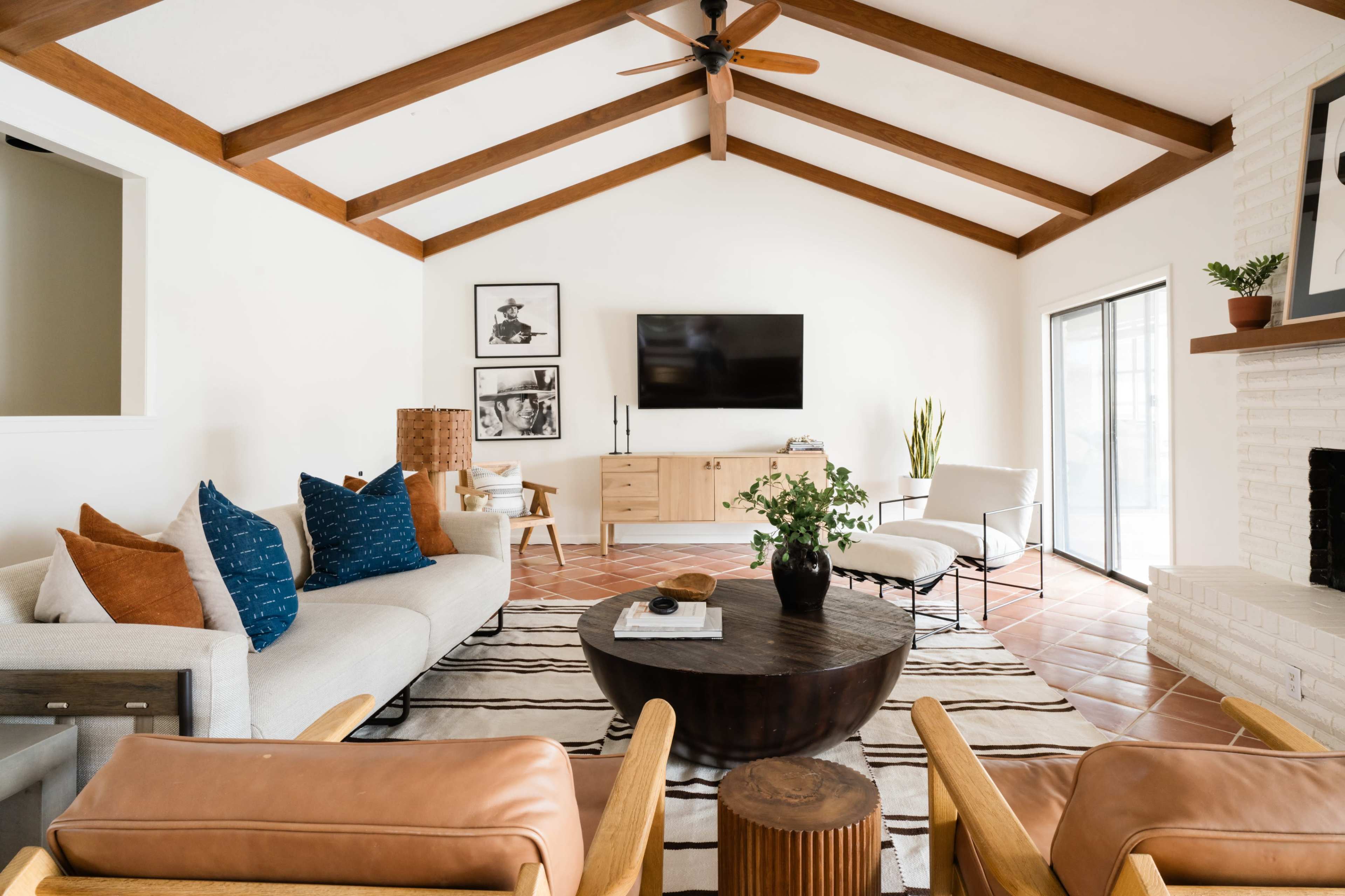The living room features a sofa with blue and orange pillows, a round coffee table, and a wall-mounted television, all set beneath a vaulted wooden ceiling.