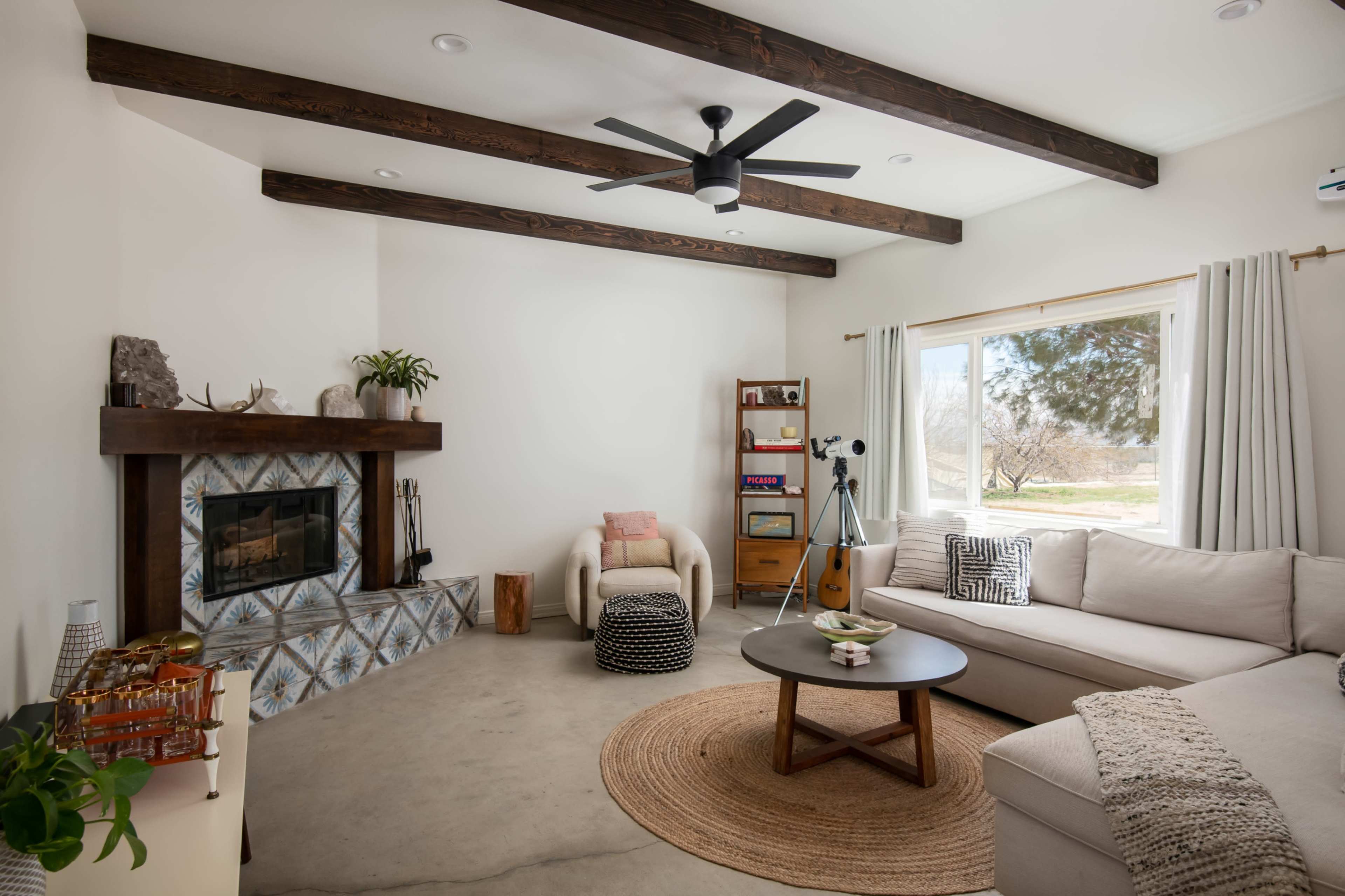 A modern living room features a light-colored couch, a round coffee table, a fireplace with a wooden mantel, and large windows overlooking a garden.