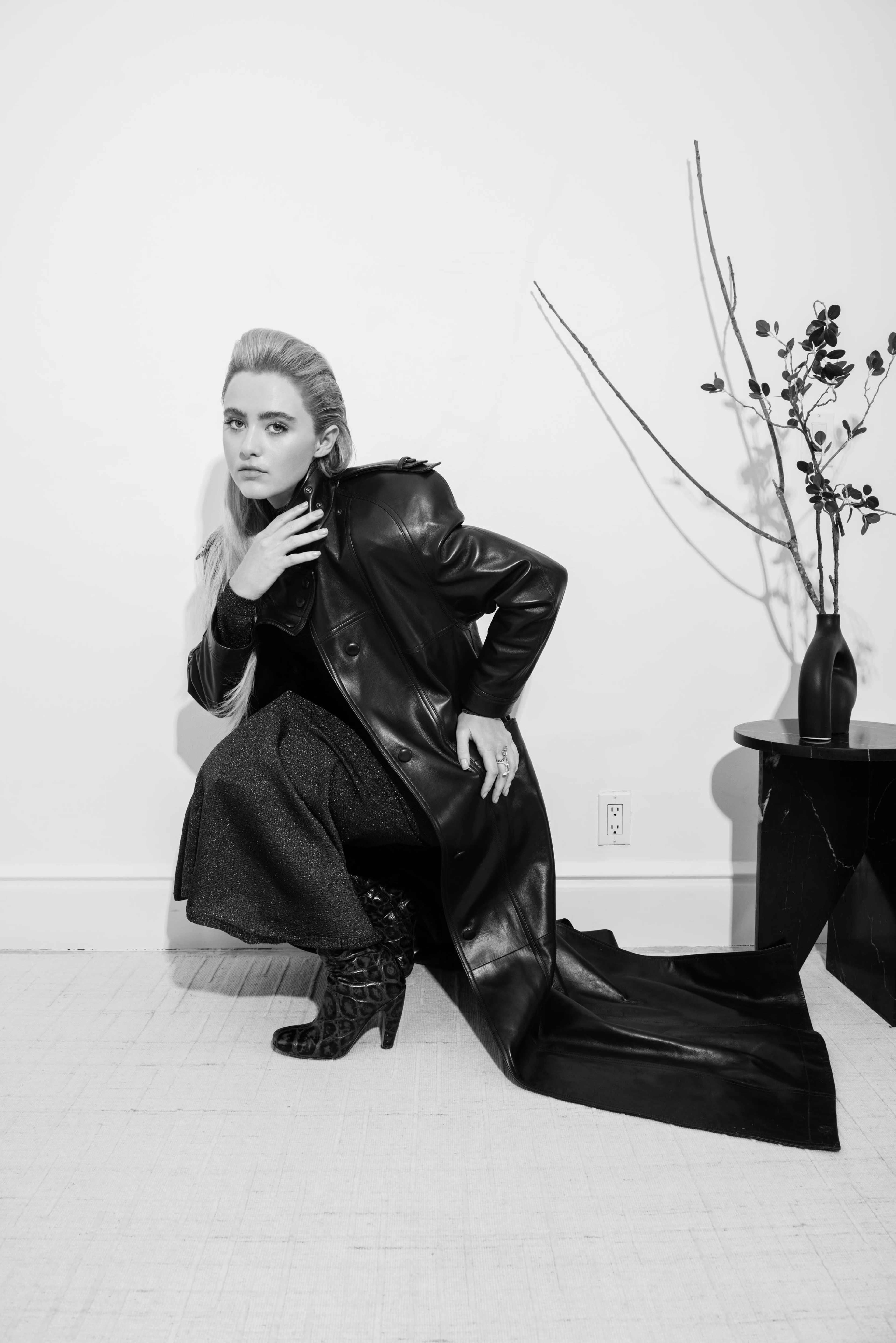 A woman in a long black leather coat and a dark skirt poses gracefully next to a minimalist black table with a vase and branches.