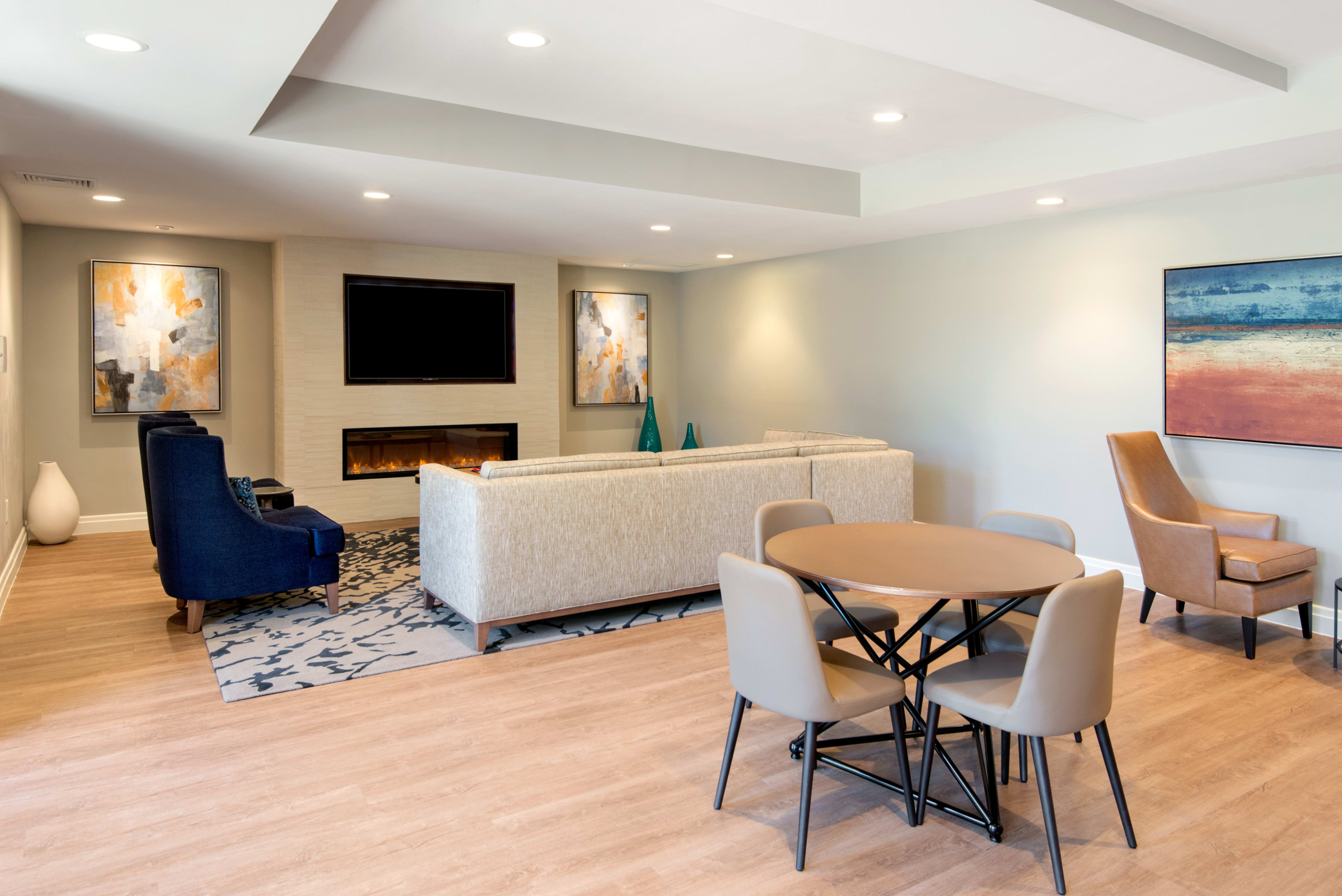 A modern living area features a sectional sofa, a round dining table with four chairs, and wall-mounted art on light-colored walls.