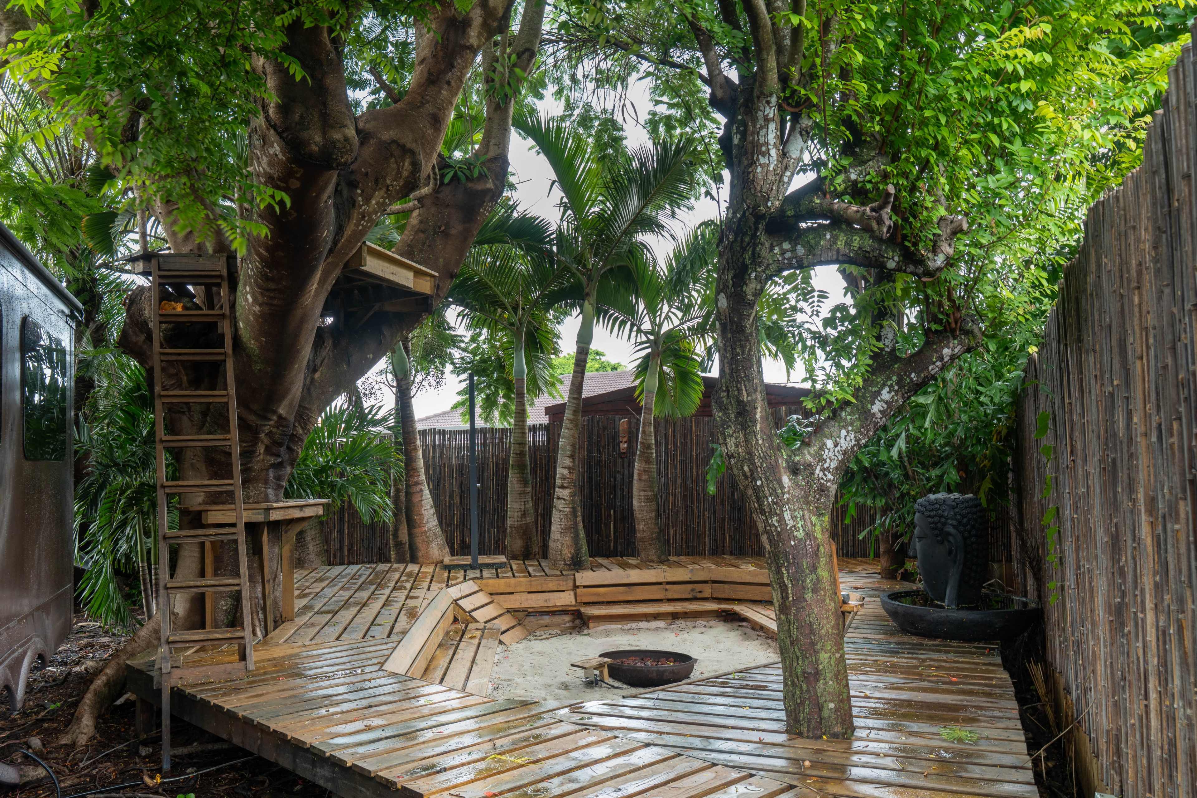 The image shows a wooden deck surrounded by trees and plants, featuring a fire pit and a small ladder leading to a platform in the branches.