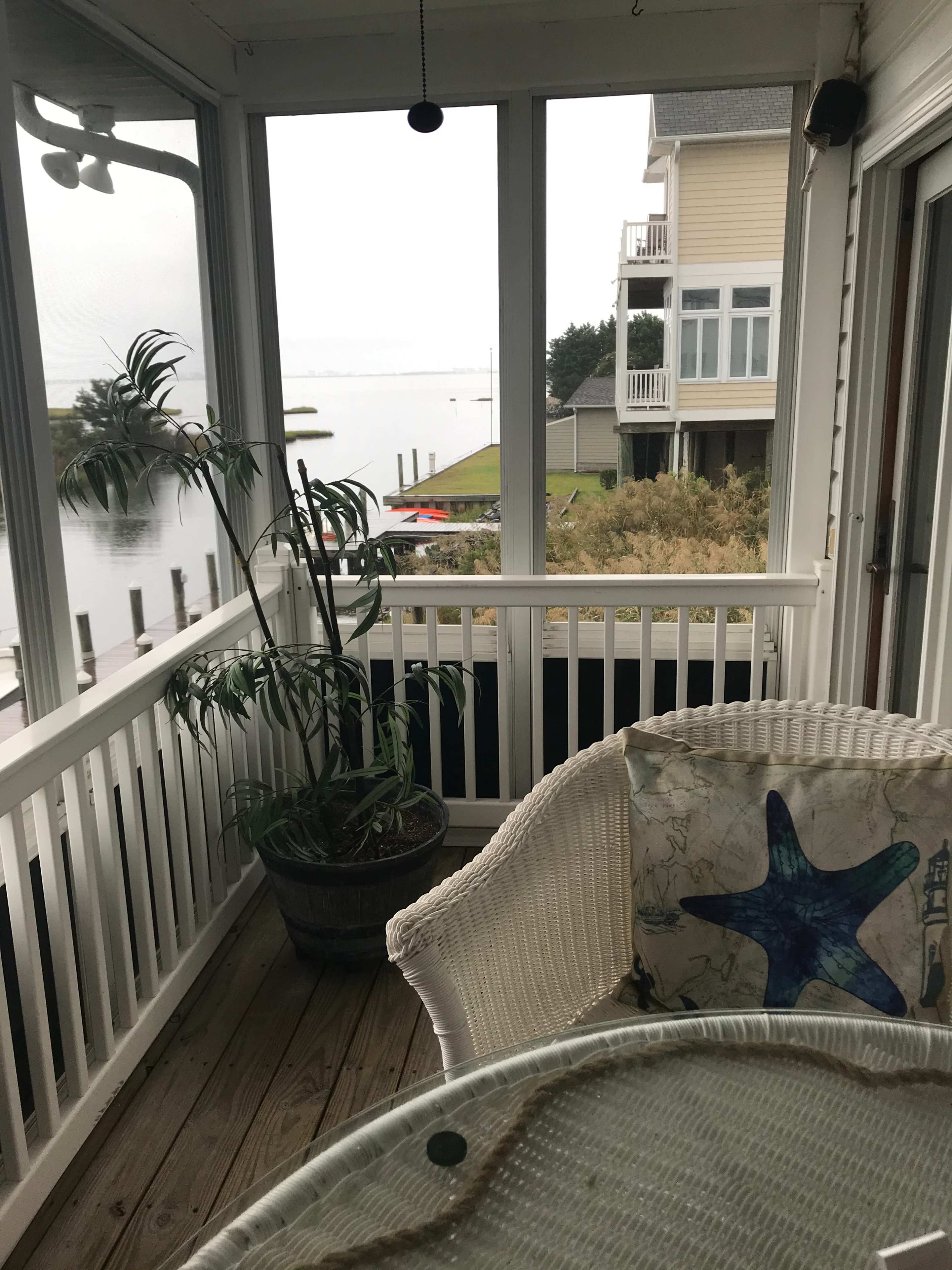 A screened-in porch features a white wicker chair with a starfish print cushion and a potted plant, overlooking a calm waterfront and nearby houses.