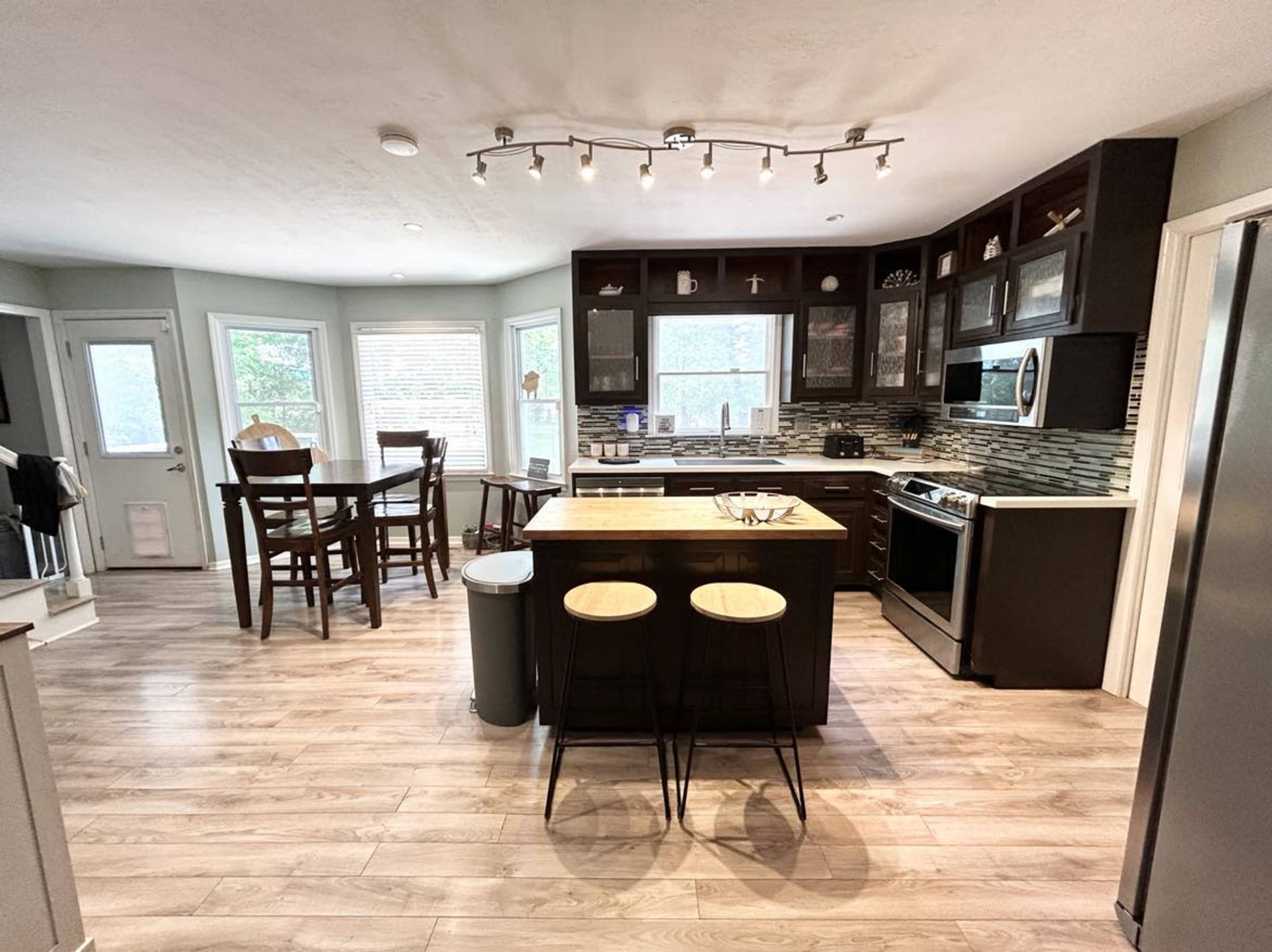 A modern kitchen with dark cabinetry, a central island with two stools, and a dining area with a table and chairs.