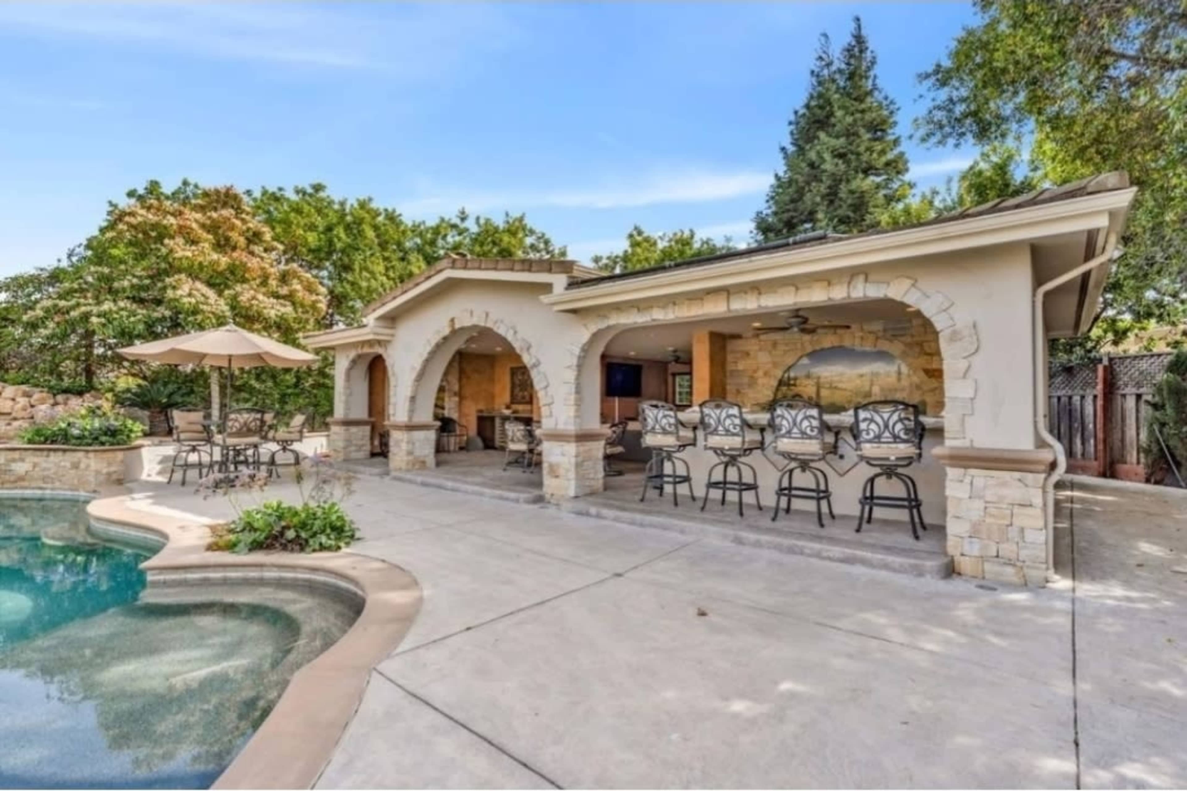 The image shows a spacious outdoor patio area with a pool, surrounded by stone arches, comfortable seating, and a shaded dining space.