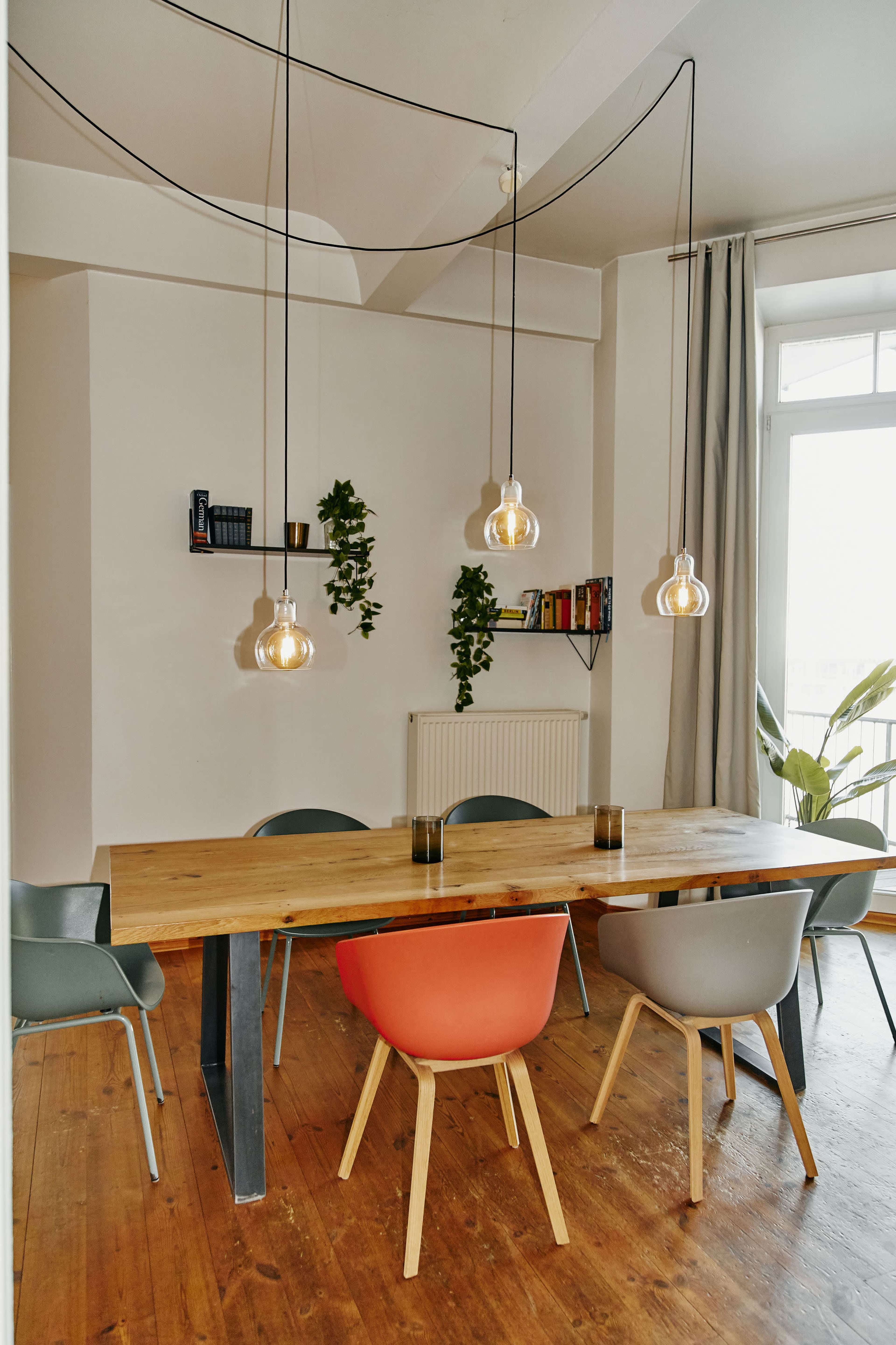 A dining area features a wooden table surrounded by colorful chairs and pendant lights, with shelves of books and greenery in the background.