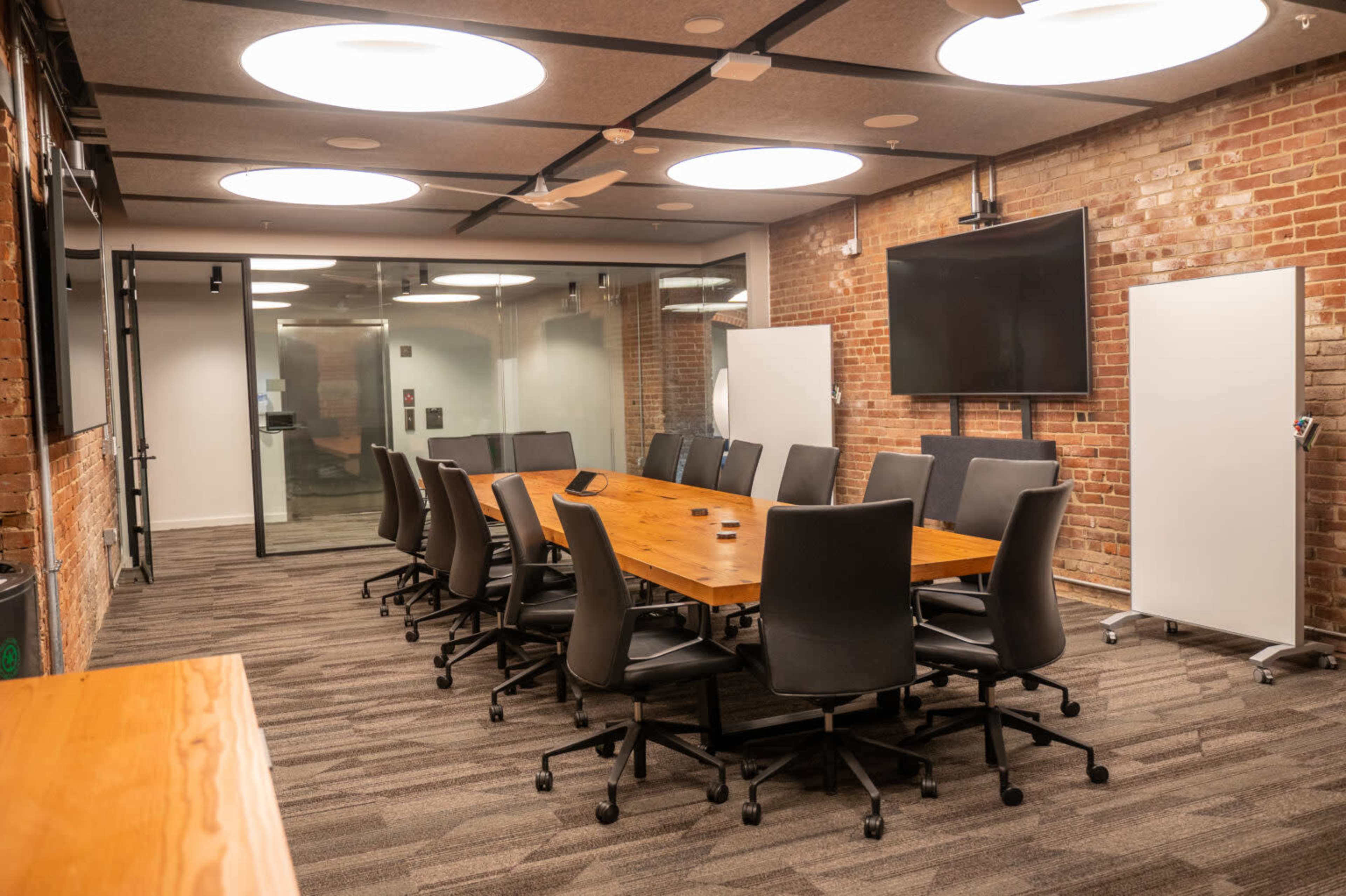 A modern conference room features a large wooden table surrounded by ergonomic chairs, with exposed brick walls and a flat-screen display.