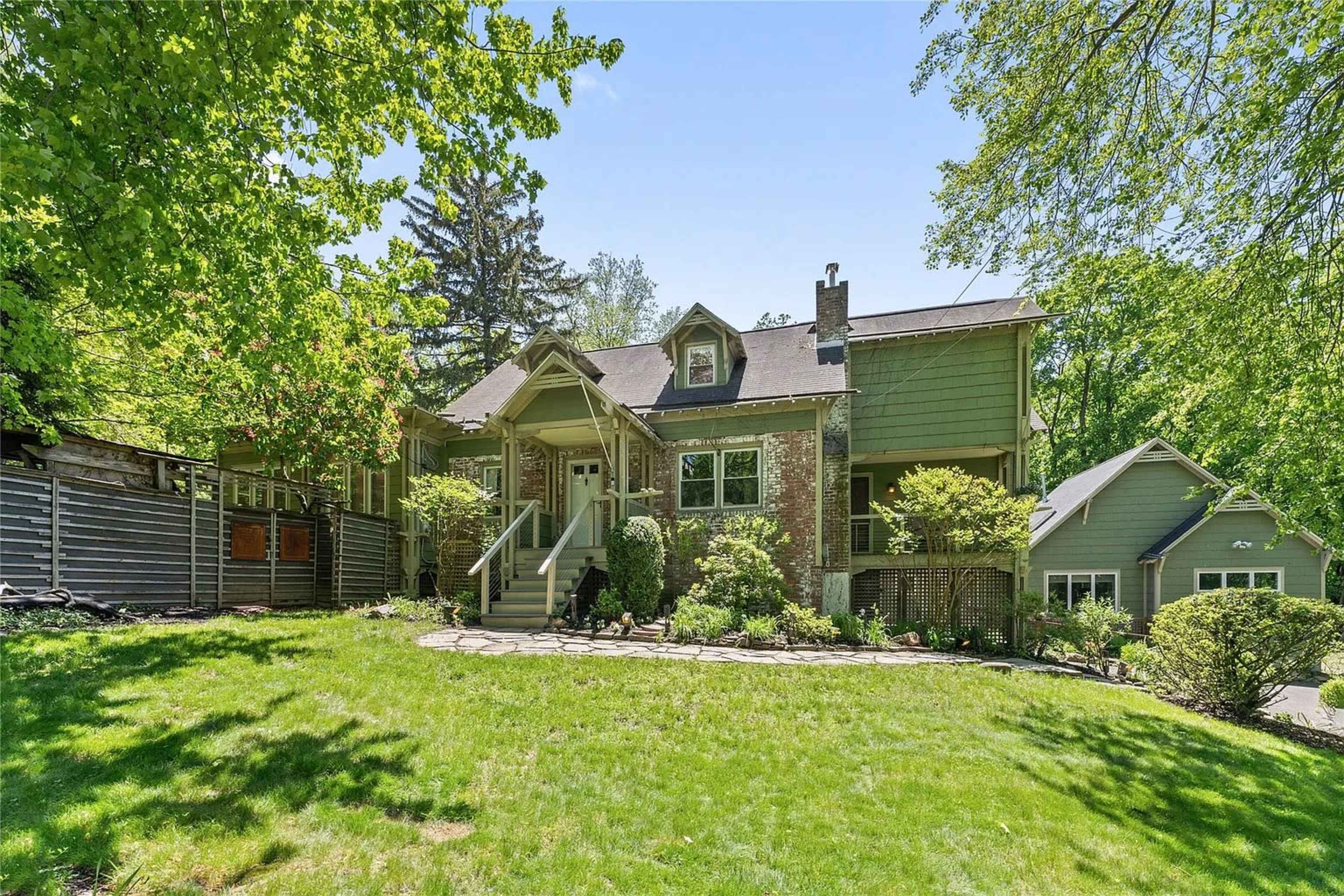 A large house with a combination of brick and green siding is surrounded by lush greenery and a well-maintained lawn.