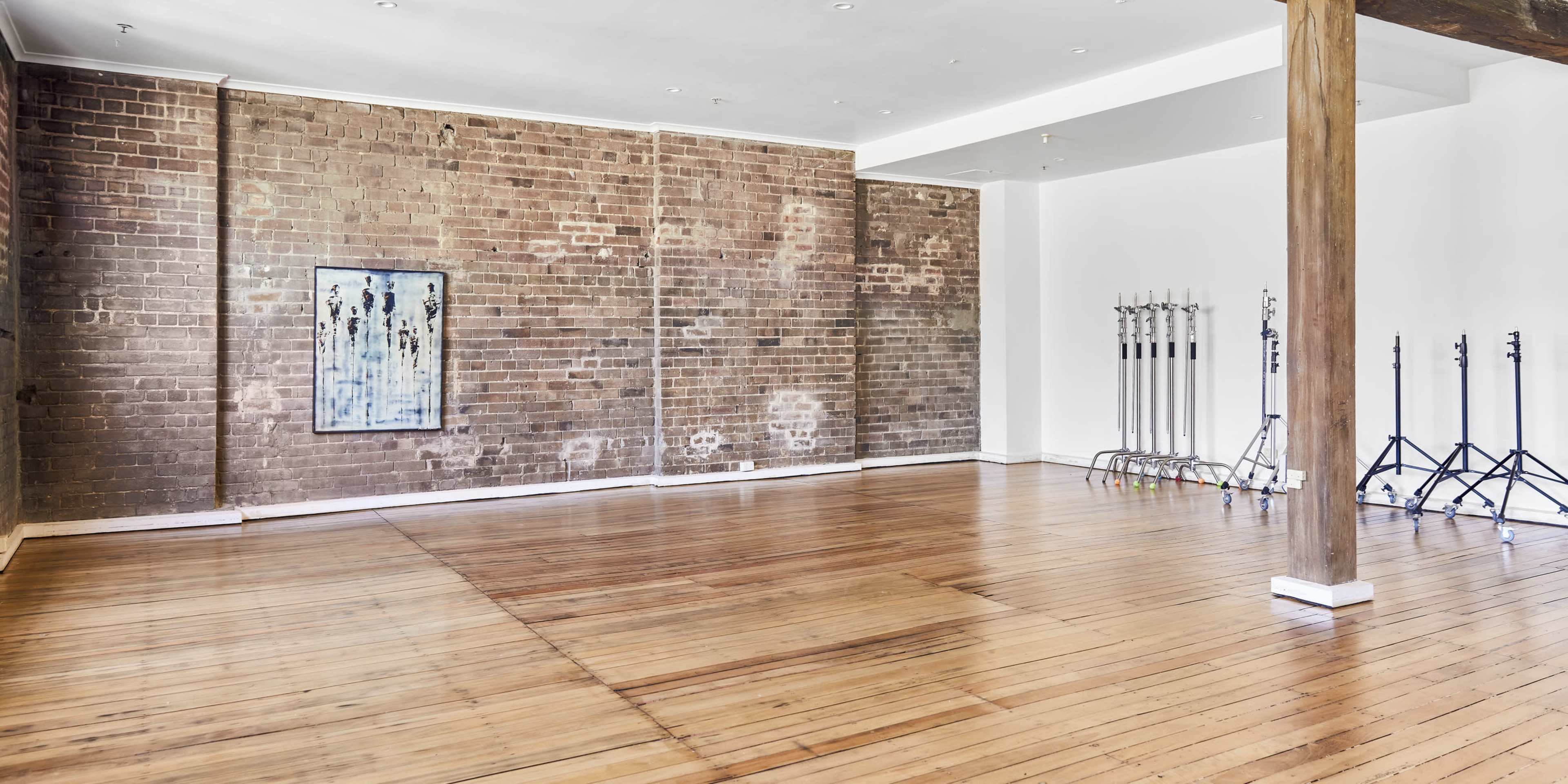 A spacious room with wooden flooring, exposed brick walls, and a few metal light stands positioned against one wall.