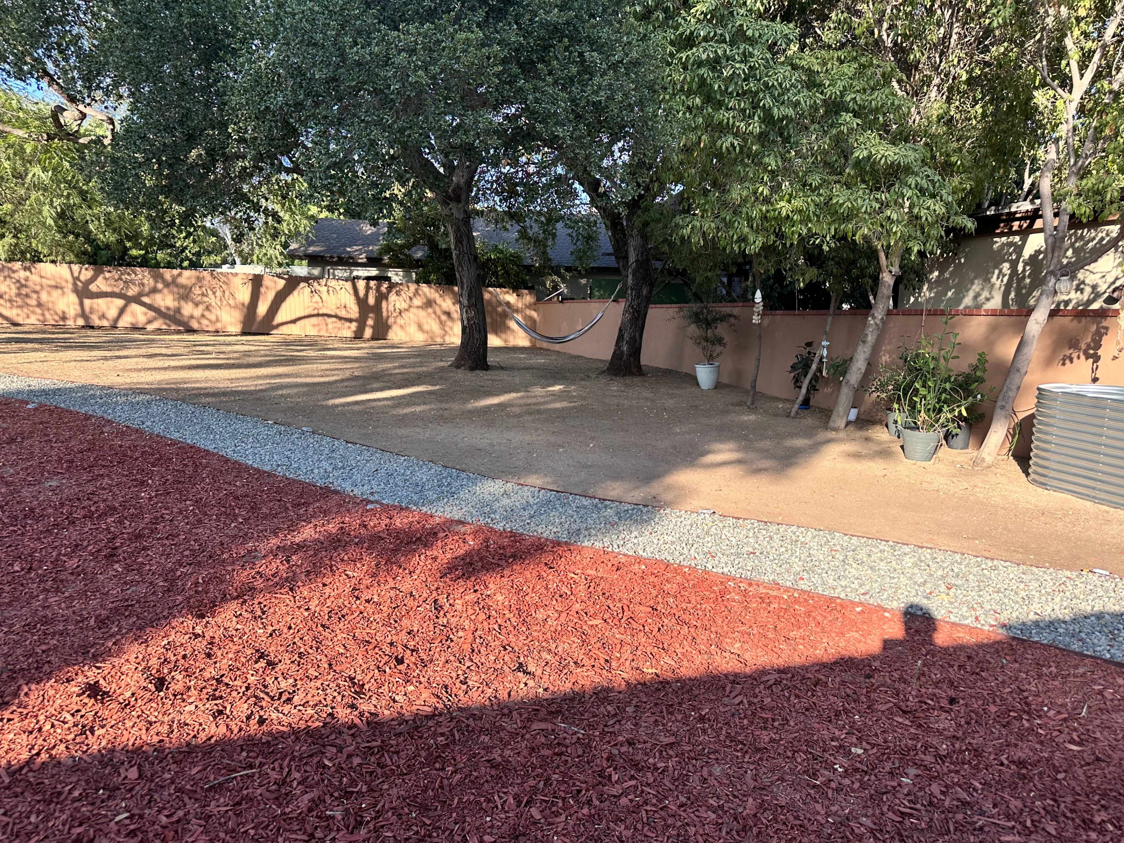 The image shows a yard with a gravel pathway, red mulch, two trees, and a hammock between them, bordered by a fence.