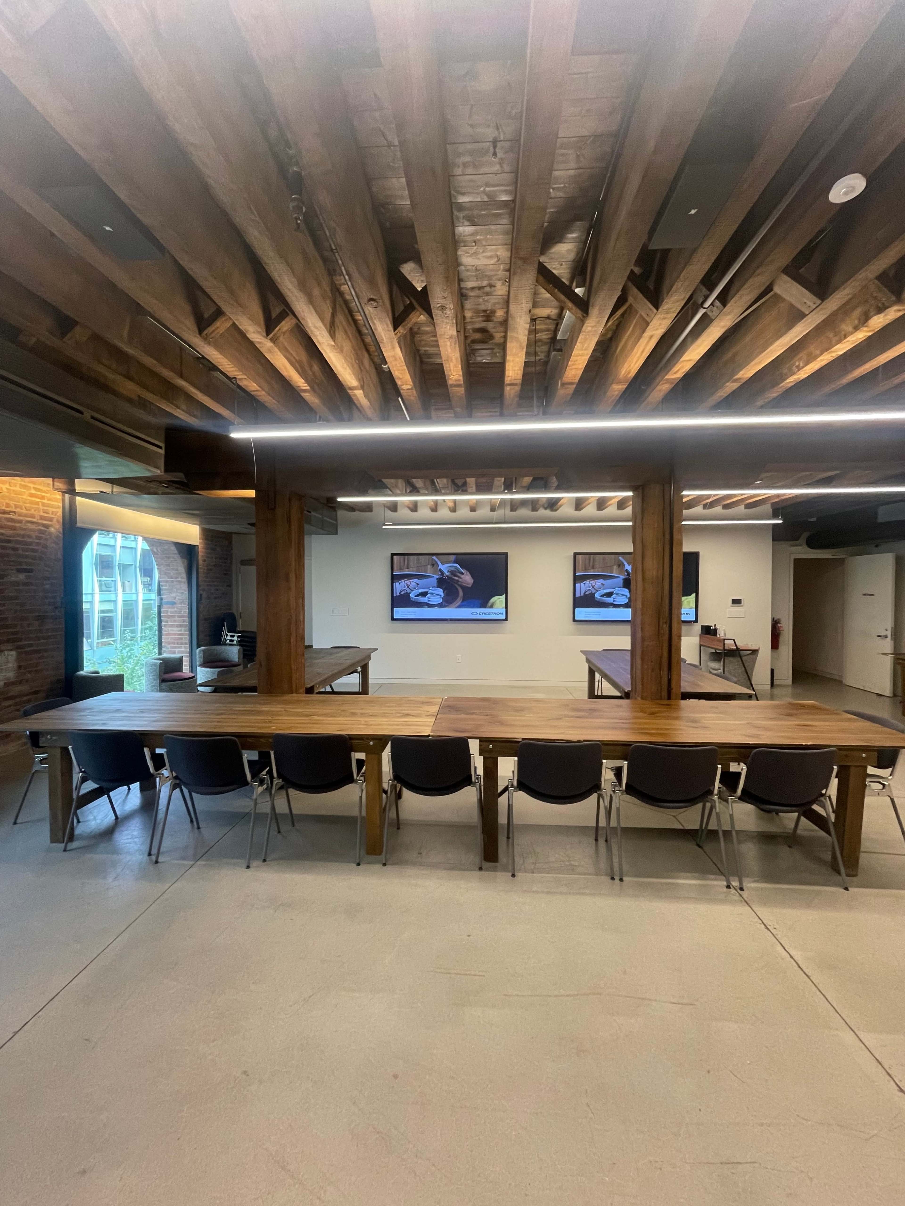 A spacious conference room features a long wooden table, several black chairs, and two screens mounted on the wall, all beneath a ceiling with exposed wooden beams.