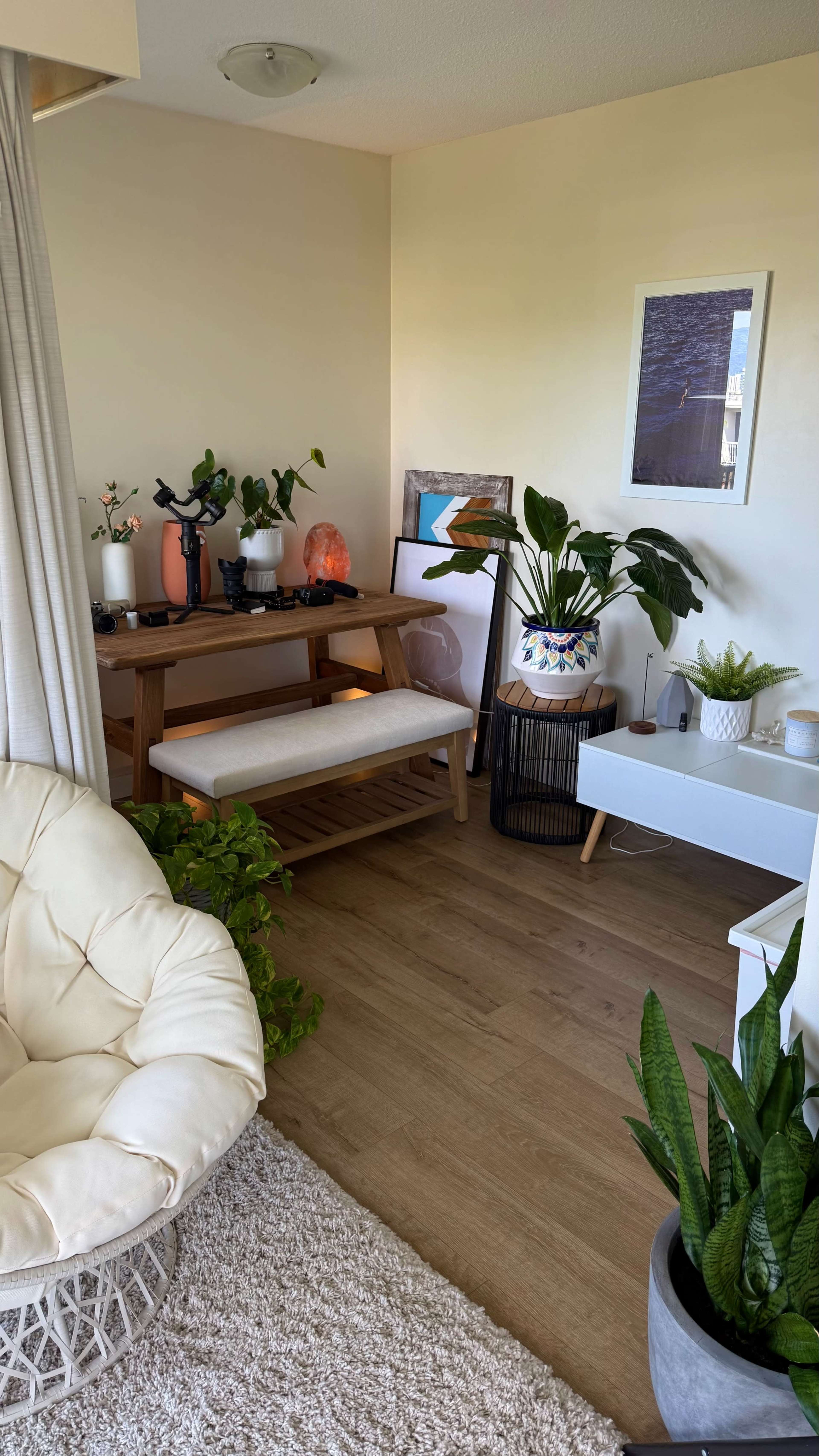 The image shows a cozy corner of a room featuring a wooden table with decorative plants, a comfortable chair, and various artworks on the walls.