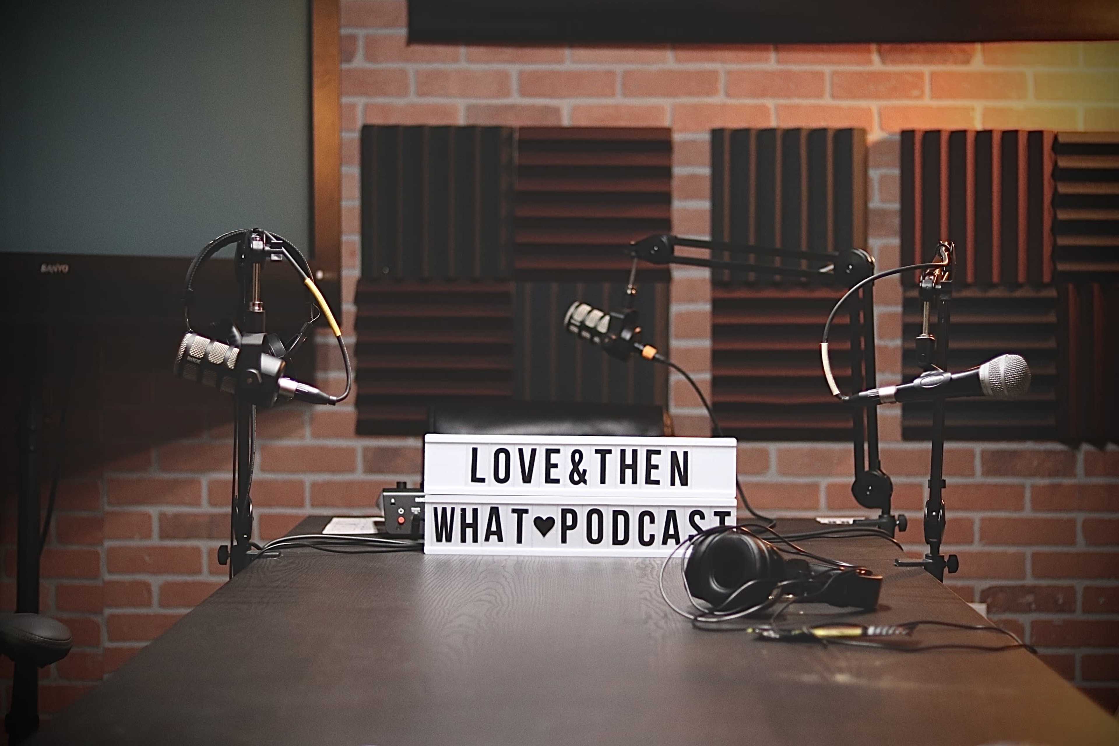 A podcast studio setup featuring two microphones, headphones, and a lightbox displaying the title "LOVE & THEN WHAT PODCAST" against a brick wall backdrop with acoustic panels.